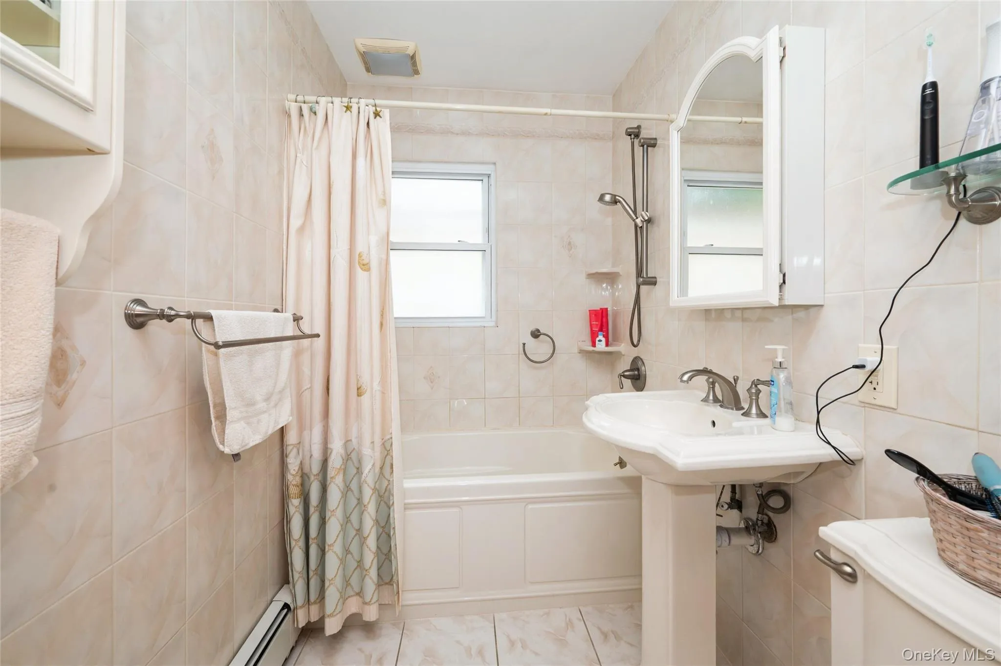 Full bathroom featuring a pedestal sink, a bathtub with an integrated shower, and light-toned wall and floor tiling Full bathroom featuring a pedestal sink, a bathtub with an integrated shower, and light-toned wall and floor tiling