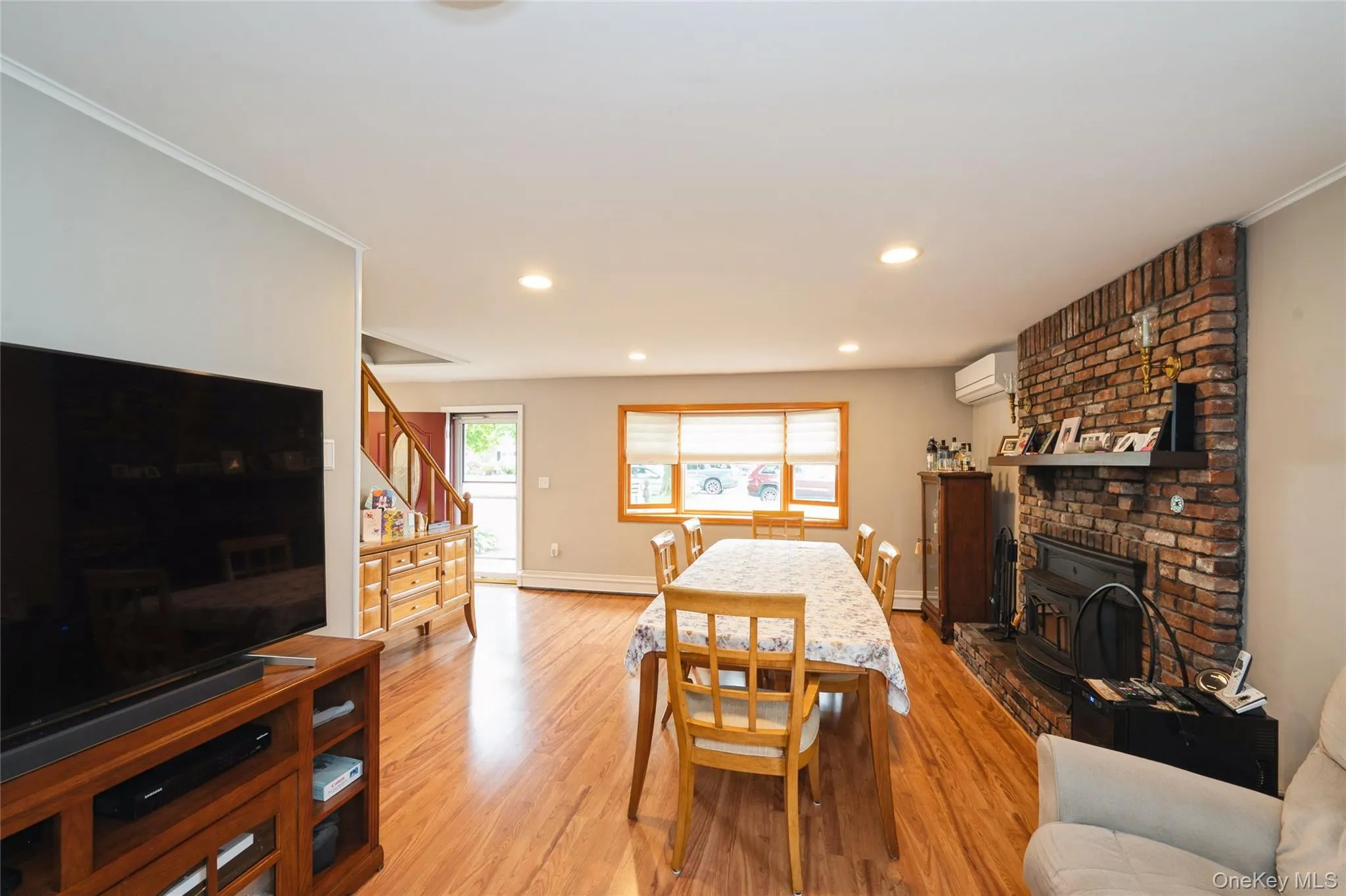Open-concept living area featuring wood-finish flooring, a brick fireplace, recessed lighting, and a window with blinds Open-concept living area featuring wood-finish flooring, a brick fireplace, recessed lighting, and a window with blinds