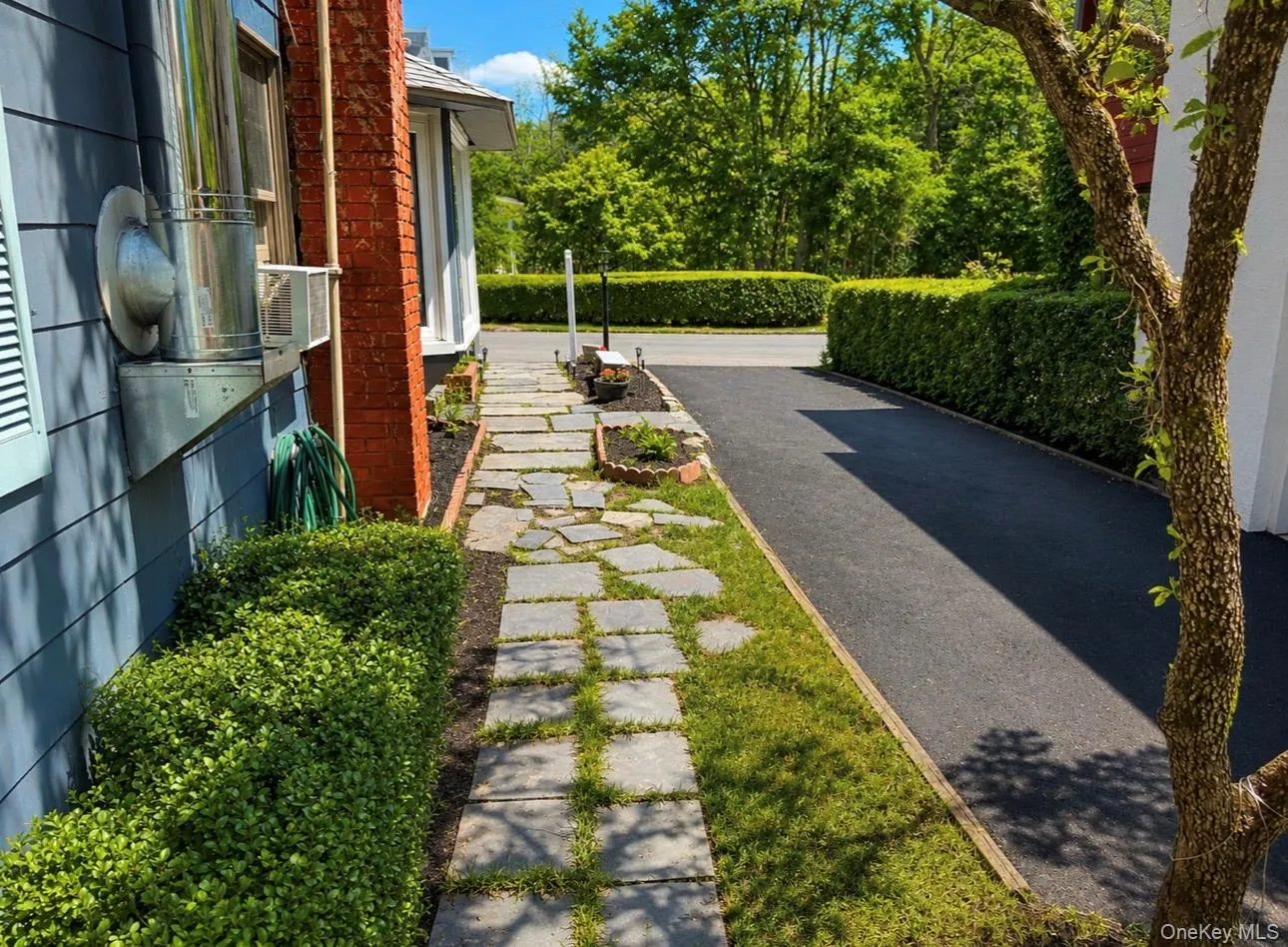 Side yard featuring an irregular stone pathway, manicured shrubs, and a paved driveway bordered by a low hedge Side yard featuring an irregular stone pathway, manicured shrubs, and a paved driveway bordered by a low hedge
