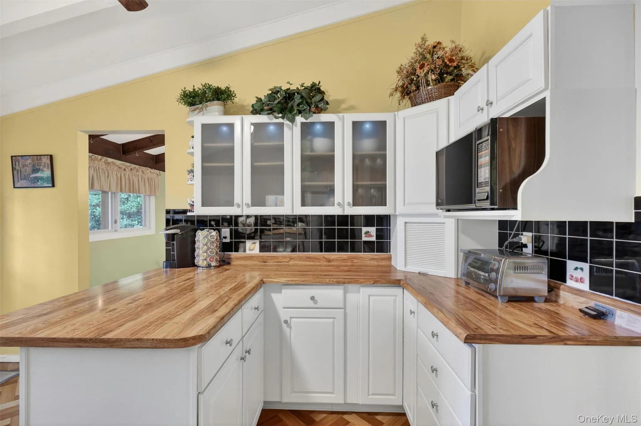Kitchen featuring vaulted ceilings with exposed beams, wood-finish flooring in a herringbone pattern, white cabinetry, breakfast bar, tile backsplash, cabinet lighting and light wood-finish countertops Kitchen featuring vaulted ceilings with exposed beams, wood-finish flooring in a herringbone pattern, white cabinetry, breakfast bar, tile backsplash, cabinet lighting and light wood-finish countertops
