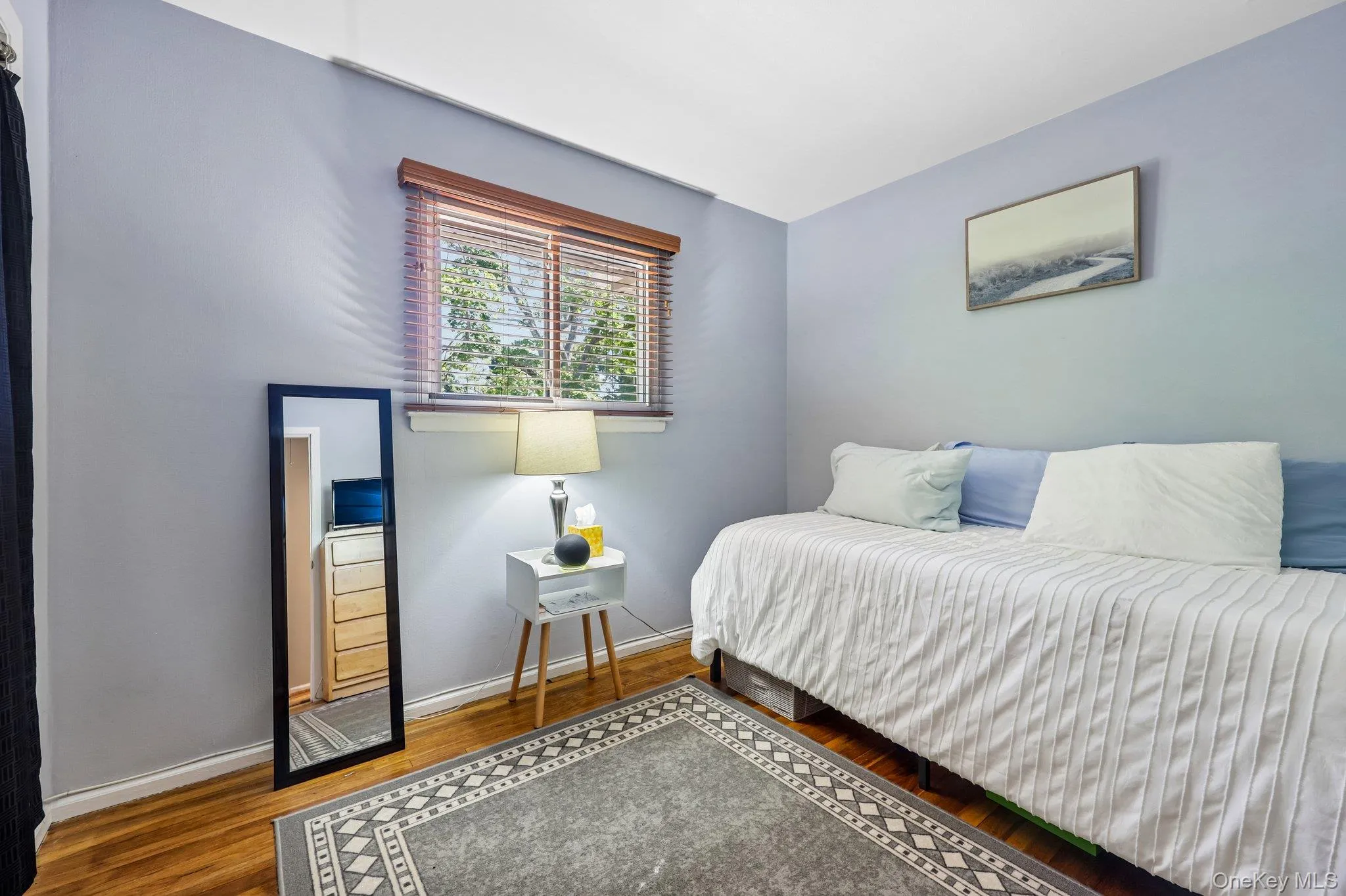 Bedroom featuring wood-finish flooring, a window with wood blinds, light-toned walls, closet, and white base and window moulding. Bedroom featuring wood-finish flooring, a window with wood blinds, light-toned walls, closet, and white base and window moulding.