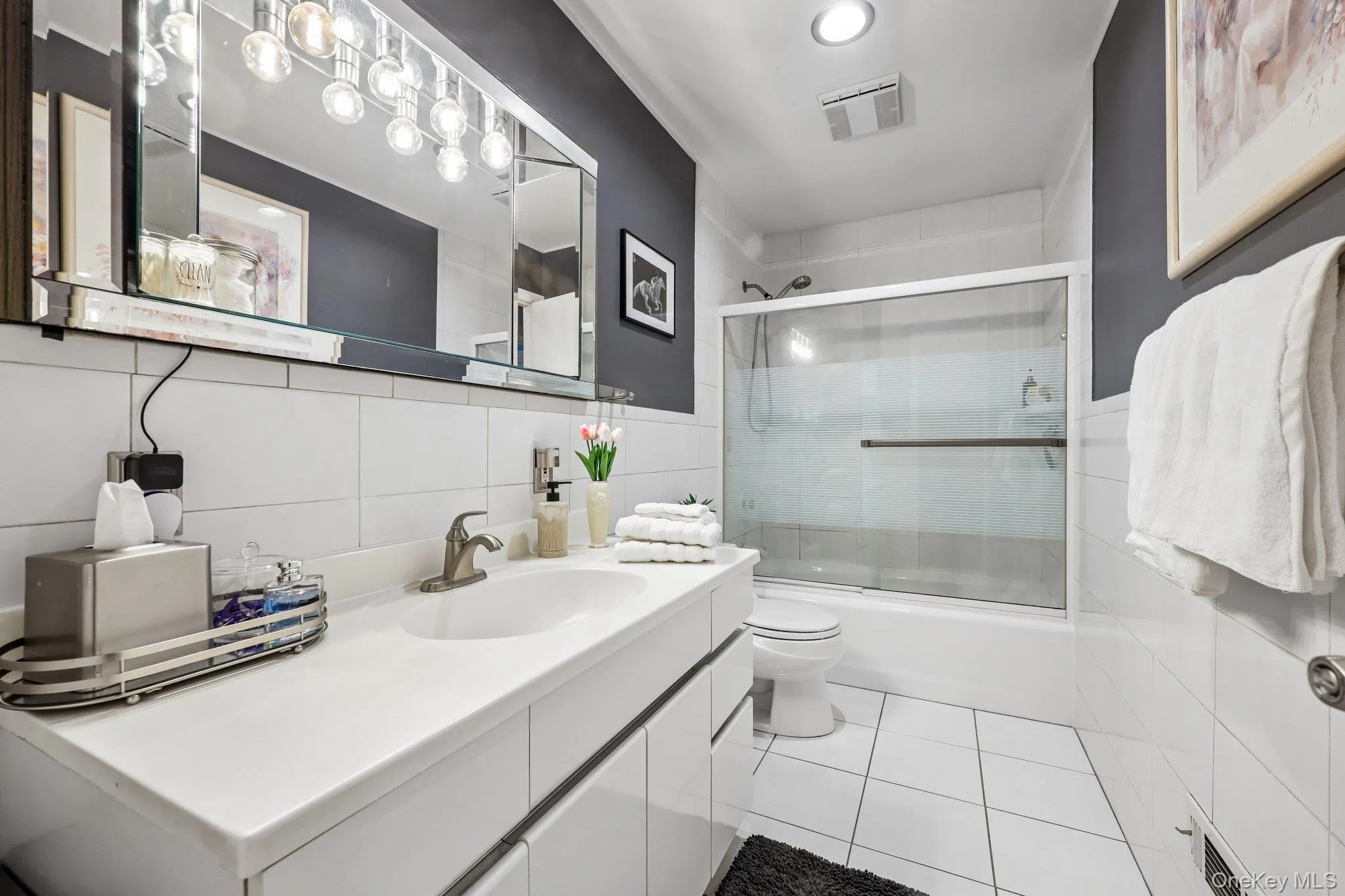 Bright White tiled bathroom featuring a large vanity with integrated sink, satin nickel modern fixtures, a large mirror with plenty of lighting, high hats, toilet, and a bathtub with a sliding glass shower door and removable shower spray. Bright White tiled bathroom featuring a large vanity with integrated sink, satin nickel modern fixtures, a large mirror with plenty of lighting, high hats, toilet, and a bathtub with a sliding glass shower door and removable shower spray.