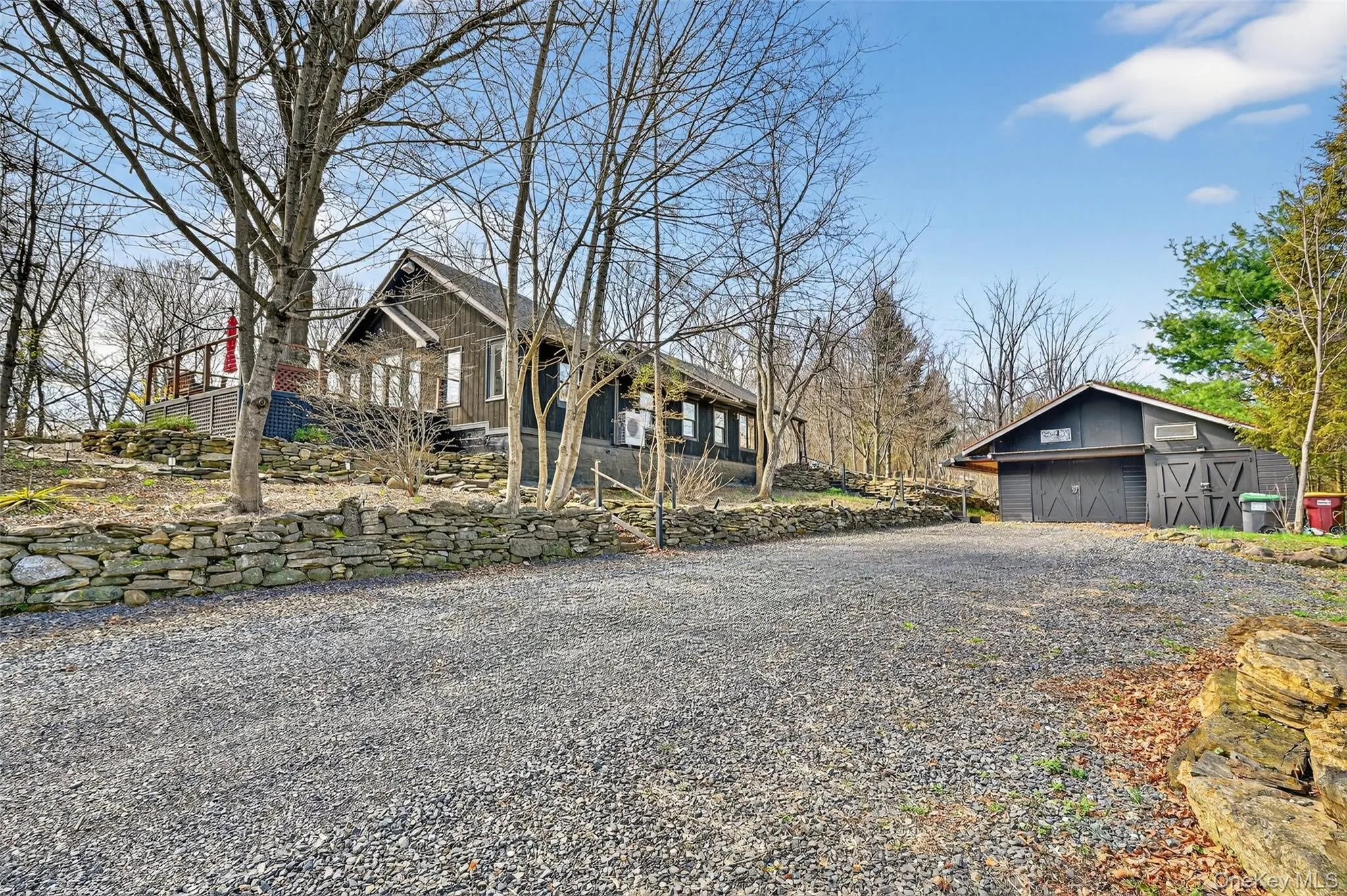 Gravel driveway servicing a property featuring a dark-toned residence with an elevated deck, a stone retaining wall, and a separate dark-toned outbuilding with two garage doors Gravel driveway servicing a property featuring a dark-toned residence with an elevated deck, a stone retaining wall, and a separate dark-toned outbuilding with two garage doors