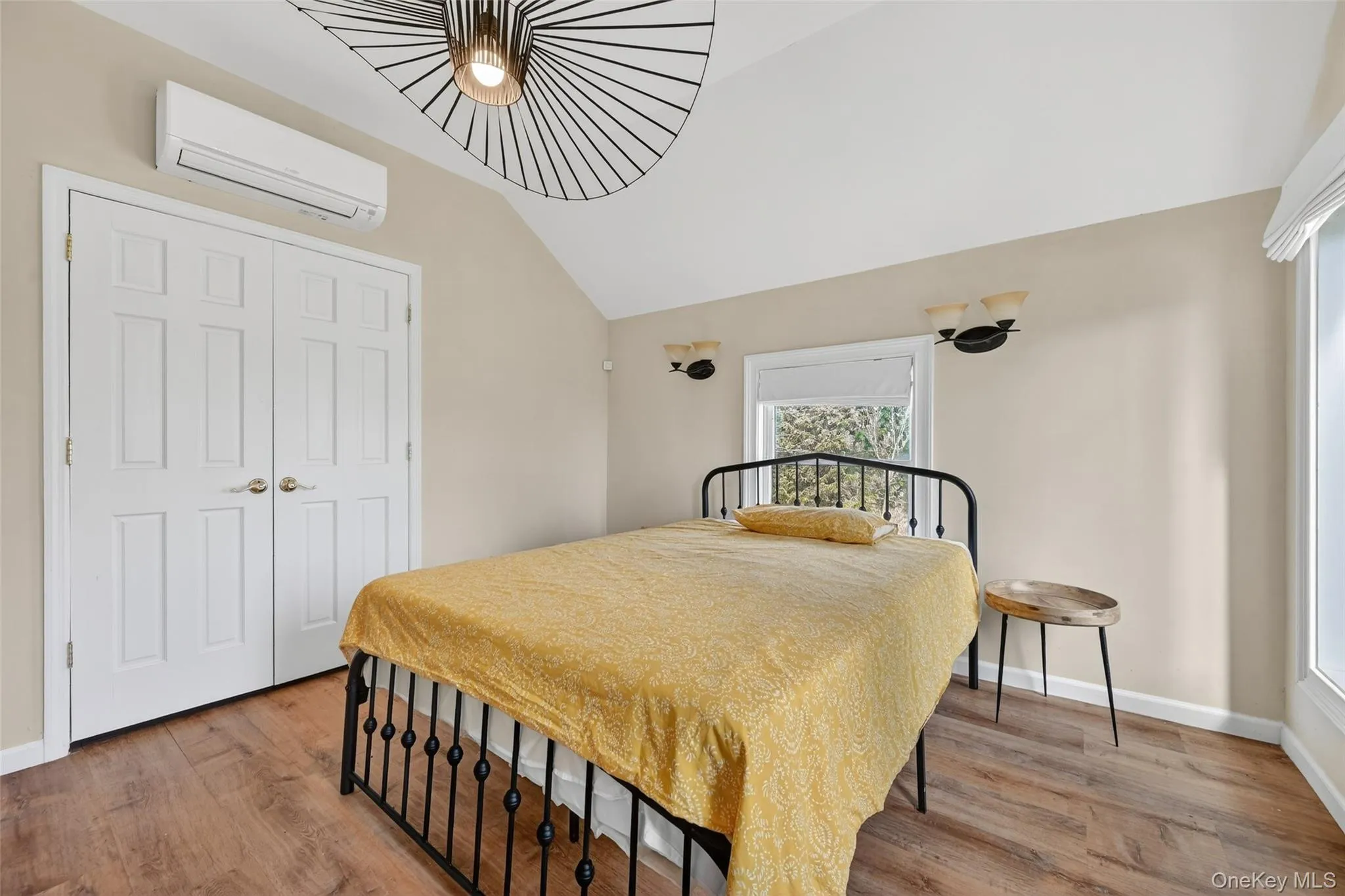 Vaulted ceiling bedroom featuring wood-finish flooring, a contemporary ceiling light fixture, bi-fold closet doors, and a wall-mounted mini-split unit Vaulted ceiling bedroom featuring wood-finish flooring, a contemporary ceiling light fixture, bi-fold closet doors, and a wall-mounted mini-split unit