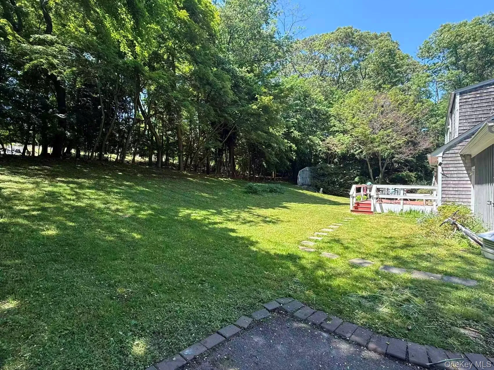 Expansive green lawn with mature perimeter trees, a stone paver path, and a rear deck with white railings Expansive green lawn with mature perimeter trees, a stone paver path, and a rear deck with white railings