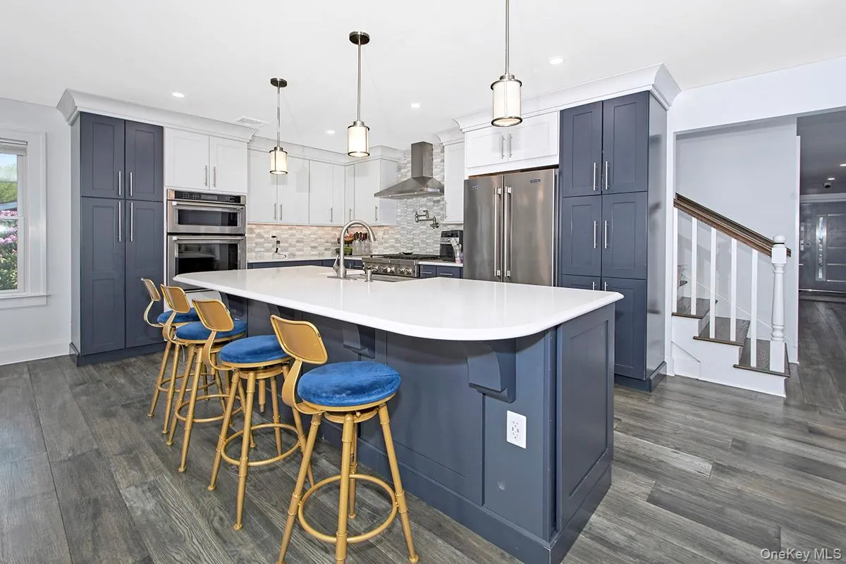 Spacious kitchen featuring a large central island with a white countertop, two-tone cabinetry with white upper and dark lower units, stainless steel double ovens, and wood-finish flooring Spacious kitchen featuring a large central island with a white countertop, two-tone cabinetry with white upper and dark lower units, stainless steel double ovens, and wood-finish flooring