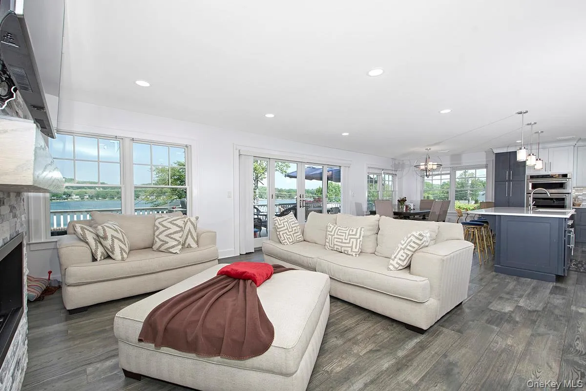 Bright open-concept living space featuring wood-finish flooring, recessed lighting, a stone-clad fireplace, and large windows with water views Bright open-concept living space featuring wood-finish flooring, recessed lighting, a stone-clad fireplace, and large windows with water views