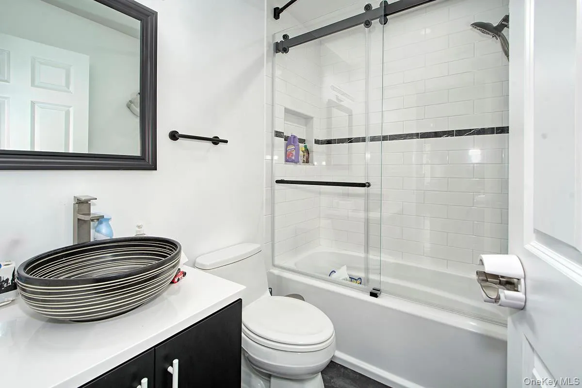 Bathroom featuring a vessel sink on a white countertop, dark wood-finish vanity with white pulls, a toilet, and a bathtub with subway tile surround and dark accent band, enclosed by a sliding glass shower door with black hardware Bathroom featuring a vessel sink on a white countertop, dark wood-finish vanity with white pulls, a toilet, and a bathtub with subway tile surround and dark accent band, enclosed by a sliding glass shower door with black hardware