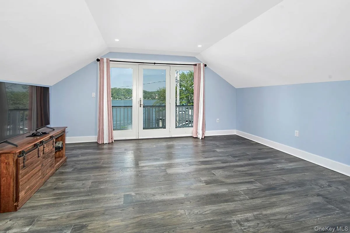 Spacious room featuring vaulted ceilings, recessed lighting, wood-finish flooring, and a double glass-panel door opening to a private balcony with water scenery Spacious room featuring vaulted ceilings, recessed lighting, wood-finish flooring, and a double glass-panel door opening to a private balcony with water scenery