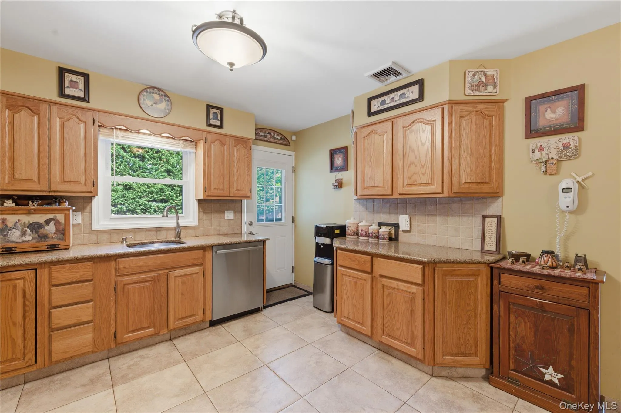 Wood cabinetry, tiled flooring, stainless steel dishwasher, a window over the sink, and a rear exterior door with a glass pane Wood cabinetry, tiled flooring, stainless steel dishwasher, a window over the sink, and a rear exterior door with a glass pane