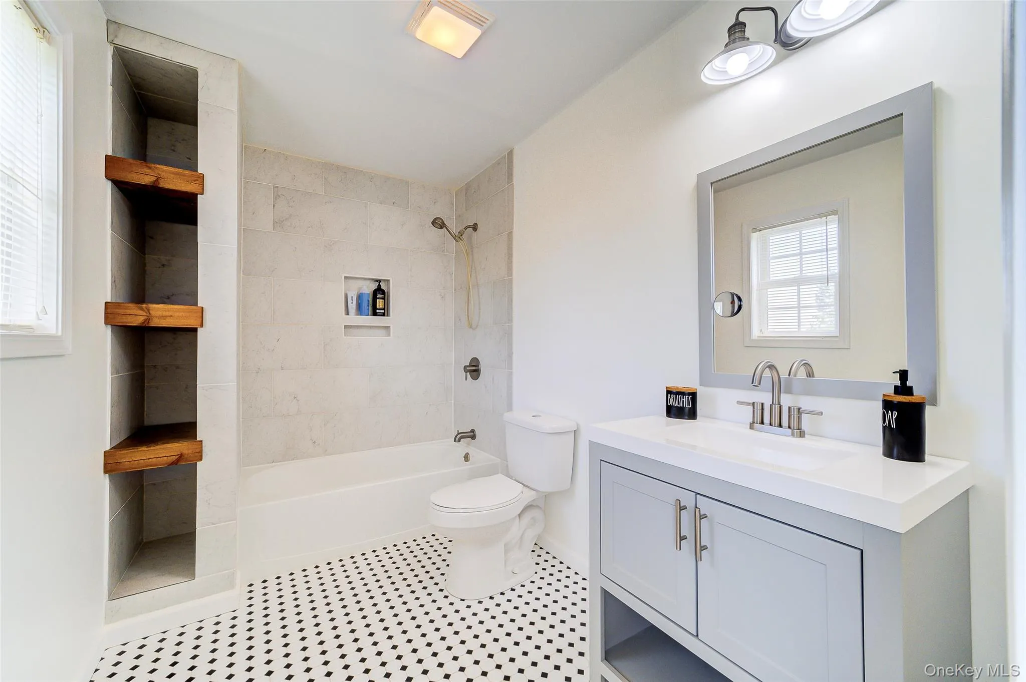 Bathroom featuring black and white hexagonal floor tiling, a light gray vanity with a white countertop, a built-in bathtub with subway tile surround, a recessed shower niche, and corner-mounted wood shelving Bathroom featuring black and white hexagonal floor tiling, a light gray vanity with a white countertop, a built-in bathtub with subway tile surround, a recessed shower niche, and corner-mounted wood shelving