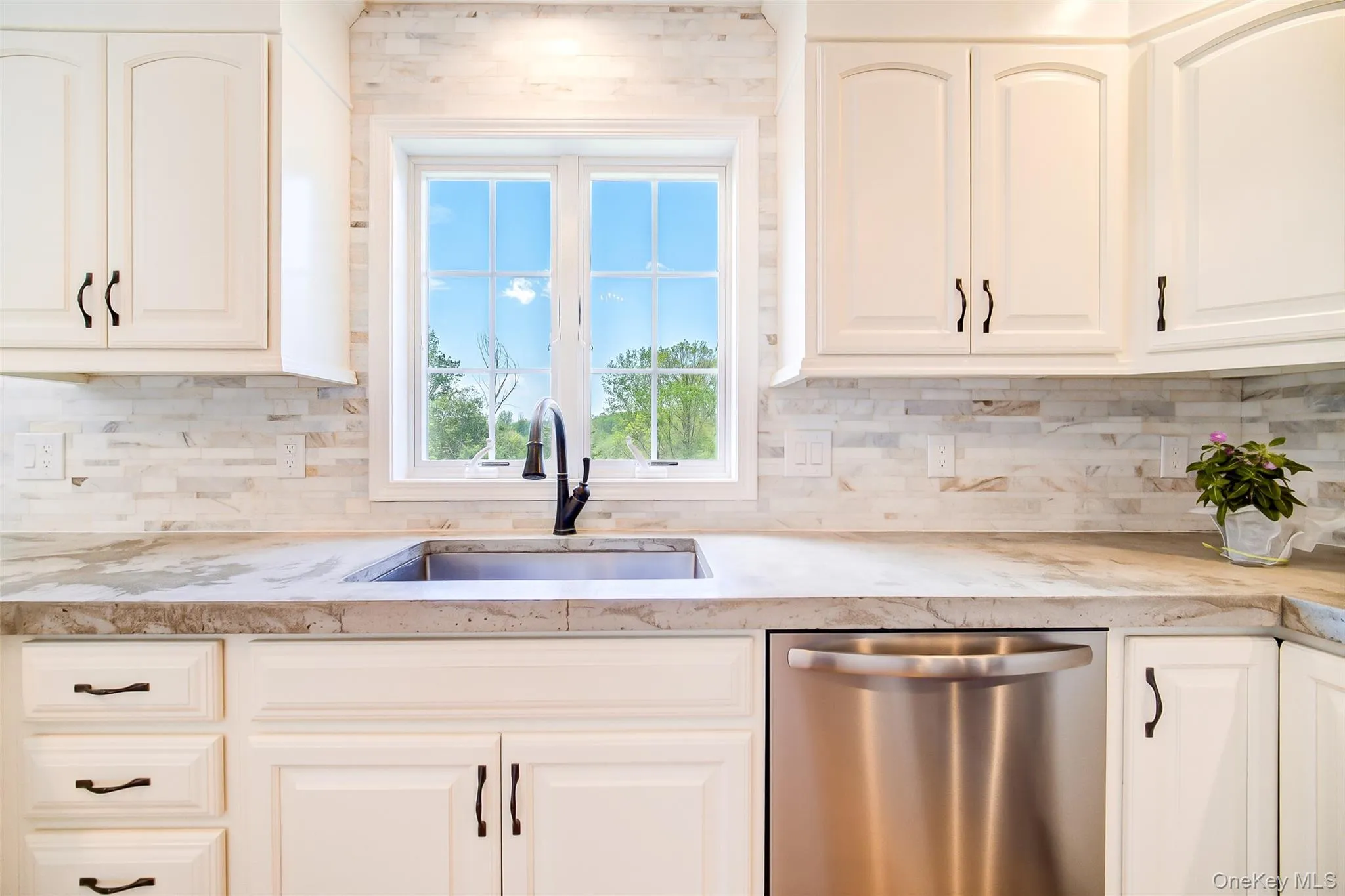 Kitchen featuring white shaker-style cabinetry, a stainless steel sink with a matte black faucet, light-toned stone countertops, and a full-height tile backsplash Kitchen featuring white shaker-style cabinetry, a stainless steel sink with a matte black faucet, light-toned stone countertops, and a full-height tile backsplash