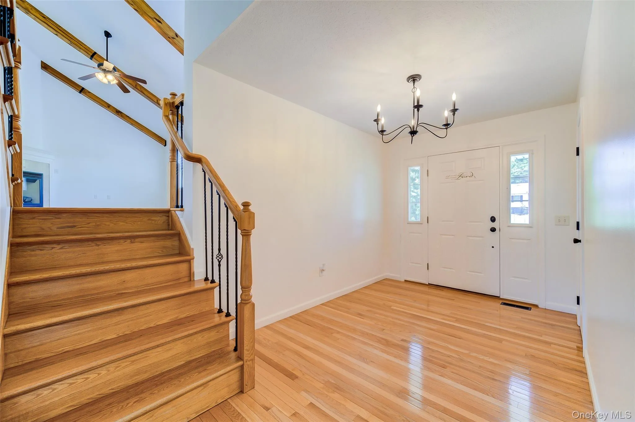 Foyer featuring natural wood-finish flooring, a white paneled entry door with sidelights, a black wrought iron chandelier, and a natural wood staircase with black metal balusters Foyer featuring natural wood-finish flooring, a white paneled entry door with sidelights, a black wrought iron chandelier, and a natural wood staircase with black metal balusters