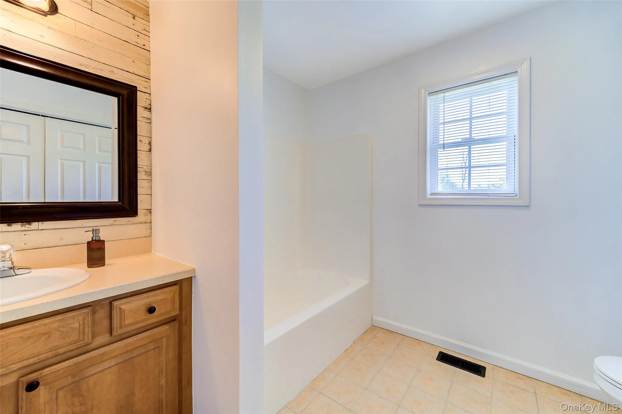 Bathroom featuring a wood-finish vanity with a light-toned countertop and integrated sink, a framed mirror, shiplap wall accent, and a white bathtub Bathroom featuring a wood-finish vanity with a light-toned countertop and integrated sink, a framed mirror, shiplap wall accent, and a white bathtub