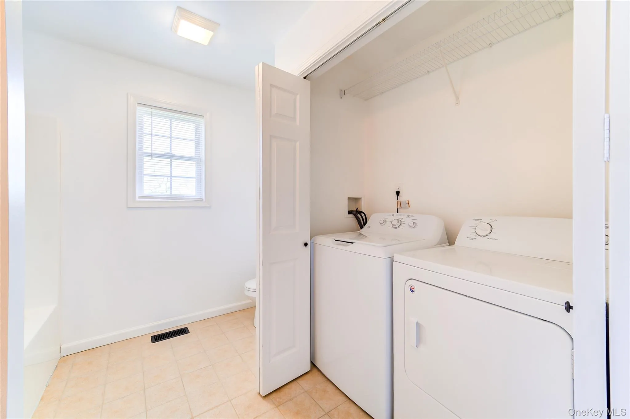 Convenient laundry area with white appliances and wire shelving, complemented by a windowed bathroom featuring a toilet and white bathtub Convenient laundry area with white appliances and wire shelving, complemented by a windowed bathroom featuring a toilet and white bathtub