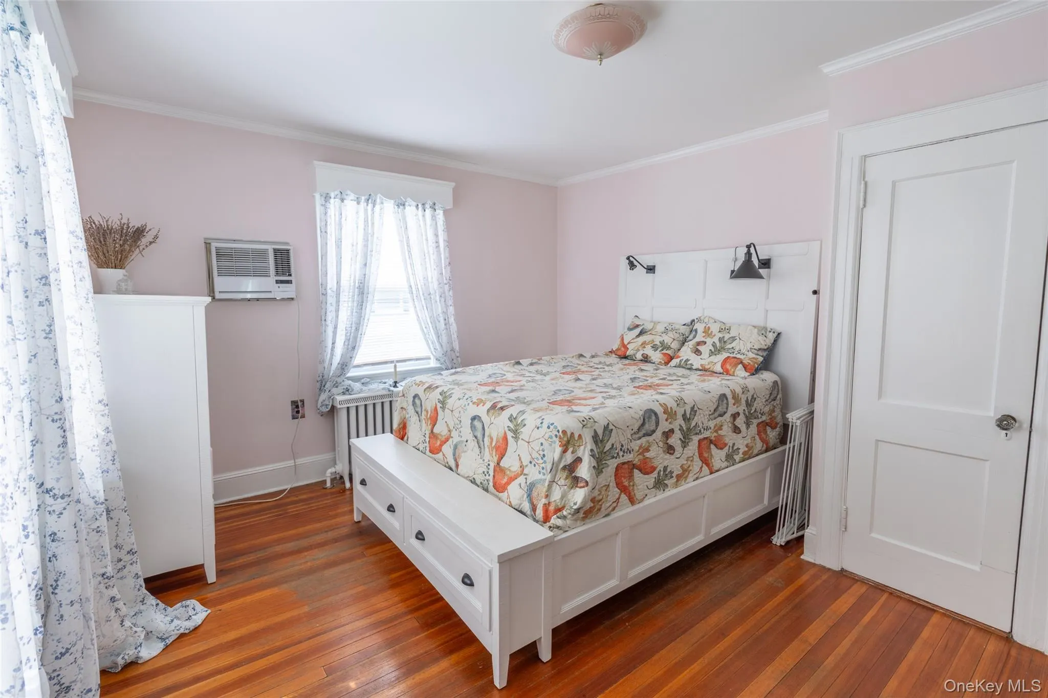 Hardwood flooring throughout bedroom featuring a window with a radiator below, a ceiling-mounted light fixture, and a white panel door Hardwood flooring throughout bedroom featuring a window with a radiator below, a ceiling-mounted light fixture, and a white panel door