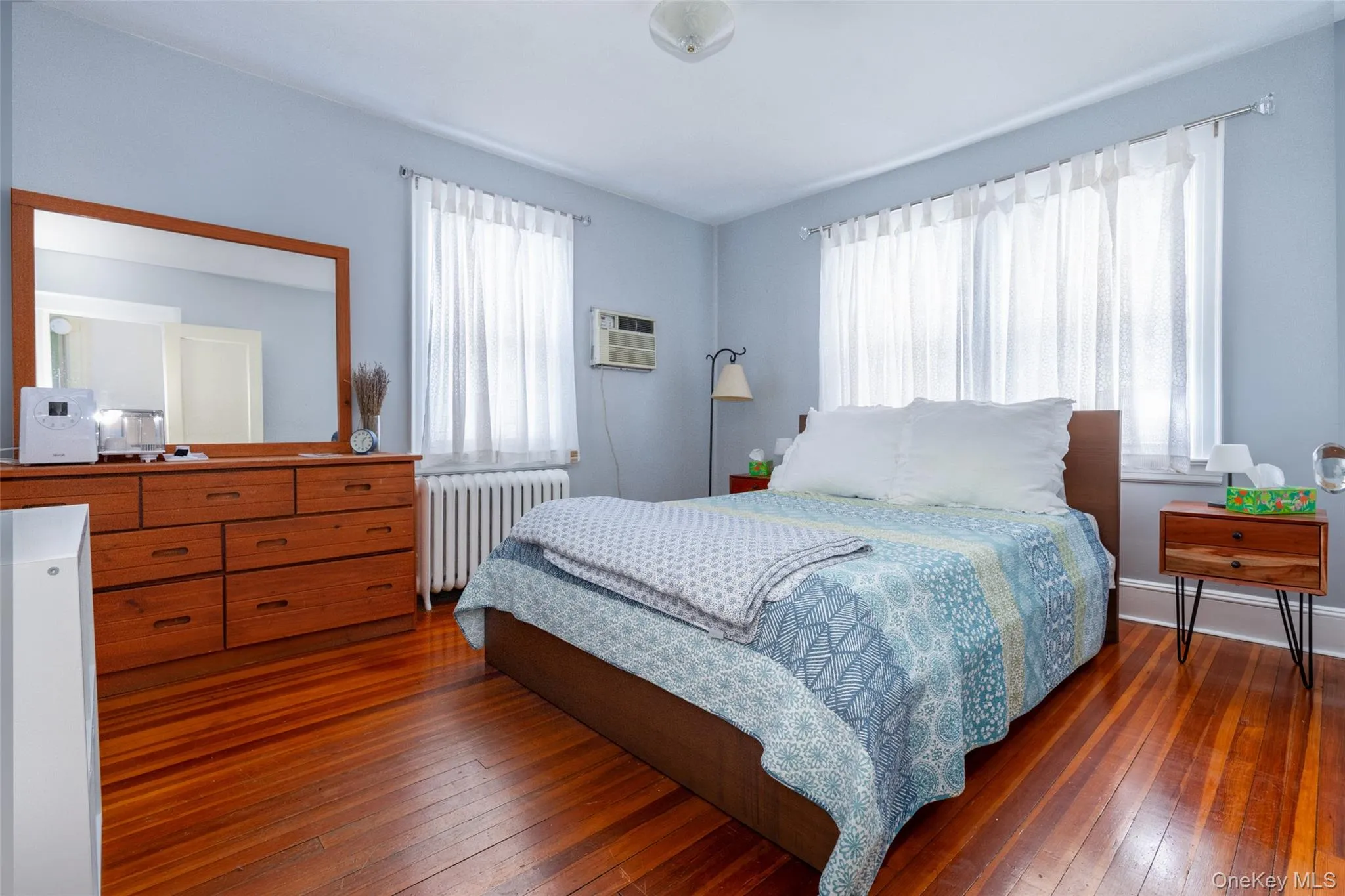 Bedroom featuring rich wood-finish flooring, light blue wall paint, and two windows providing natural illumination Bedroom featuring rich wood-finish flooring, light blue wall paint, and two windows providing natural illumination