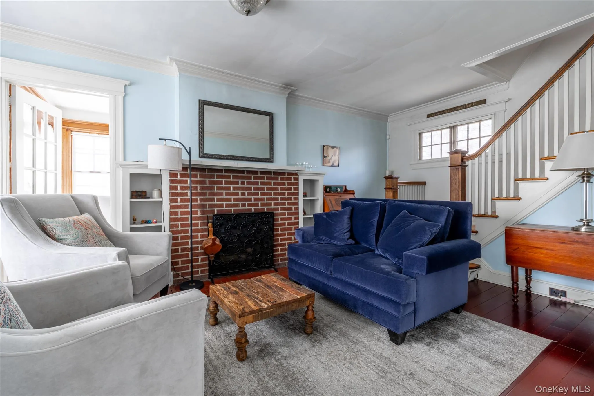 Living area featuring a brick fireplace with a white mantel and built-in shelving, hardwood flooring, and a staircase with wood newel posts and handrail Living area featuring a brick fireplace with a white mantel and built-in shelving, hardwood flooring, and a staircase with wood newel posts and handrail