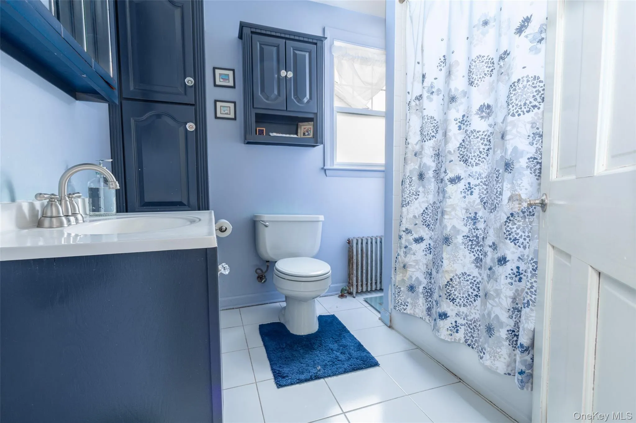 Full bathroom featuring a dark blue vanity with integrated sink, coordinating dark blue cabinetry, white square tile flooring, a white toilet, and a window for natural light Full bathroom featuring a dark blue vanity with integrated sink, coordinating dark blue cabinetry, white square tile flooring, a white toilet, and a window for natural light