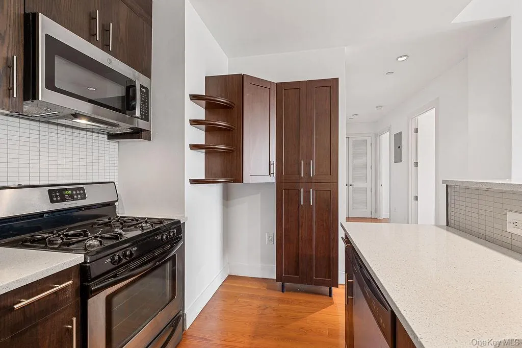 Kitchen featuring stainless steel appliances, dark wood-finish cabinetry, light-toned countertops, a white subway tile backsplash, and wood-finish flooring Kitchen featuring stainless steel appliances, dark wood-finish cabinetry, light-toned countertops, a white subway tile backsplash, and wood-finish flooring