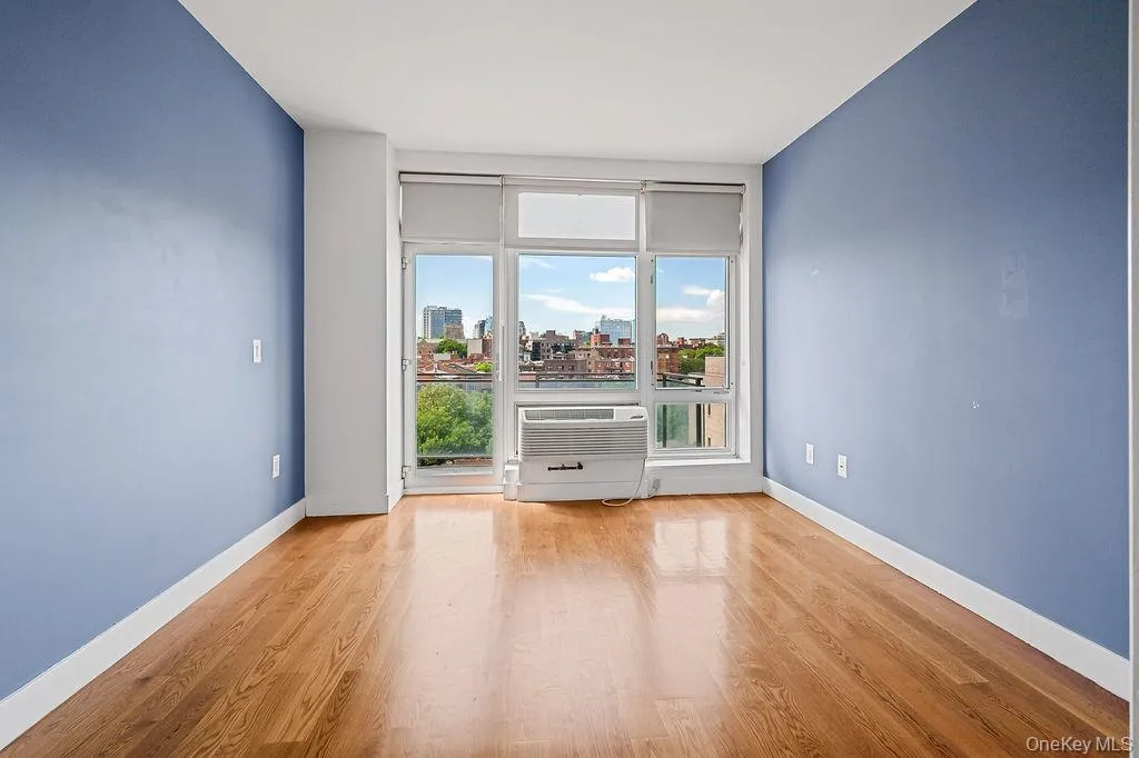 Bright room featuring wood-finish flooring, a large window with white framing, and a wall-mounted air conditioning unit Bright room featuring wood-finish flooring, a large window with white framing, and a wall-mounted air conditioning unit