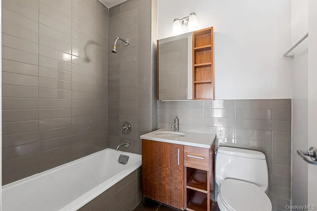Bathroom featuring a bathtub with overhead shower, grey subway tile surround, wood-finish vanity with countertop sink, wall-mounted mirror with integrated shelving, and white toilet Bathroom featuring a bathtub with overhead shower, grey subway tile surround, wood-finish vanity with countertop sink, wall-mounted mirror with integrated shelving, and white toilet