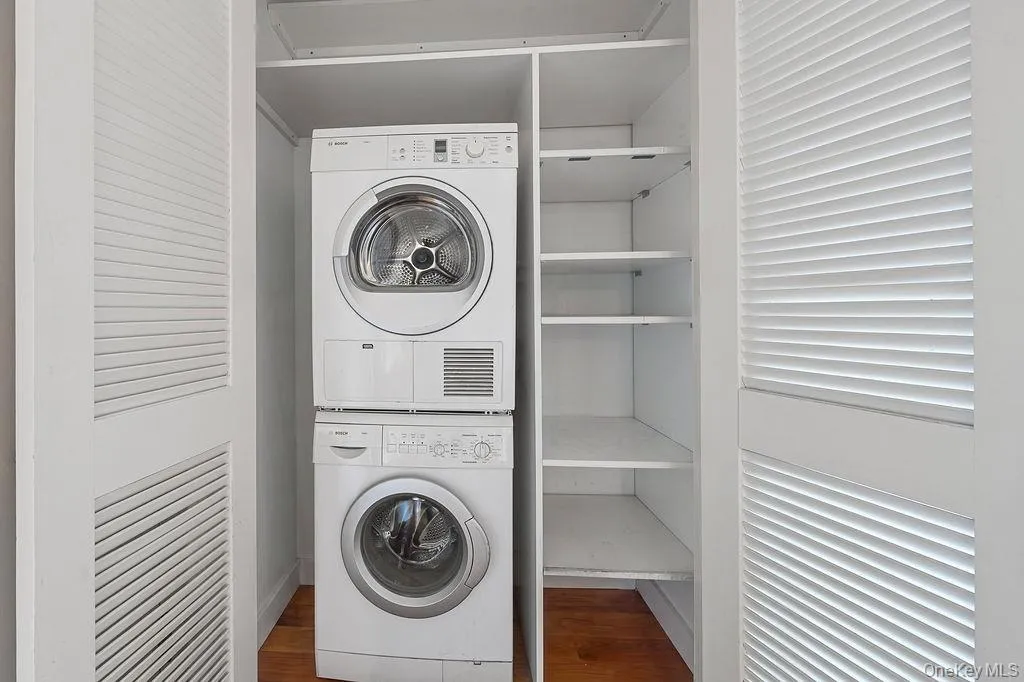 Dedicated laundry closet featuring a stacked washer and dryer, louvered bi-fold doors, integrated shelving, and hardwood flooring Dedicated laundry closet featuring a stacked washer and dryer, louvered bi-fold doors, integrated shelving, and hardwood flooring