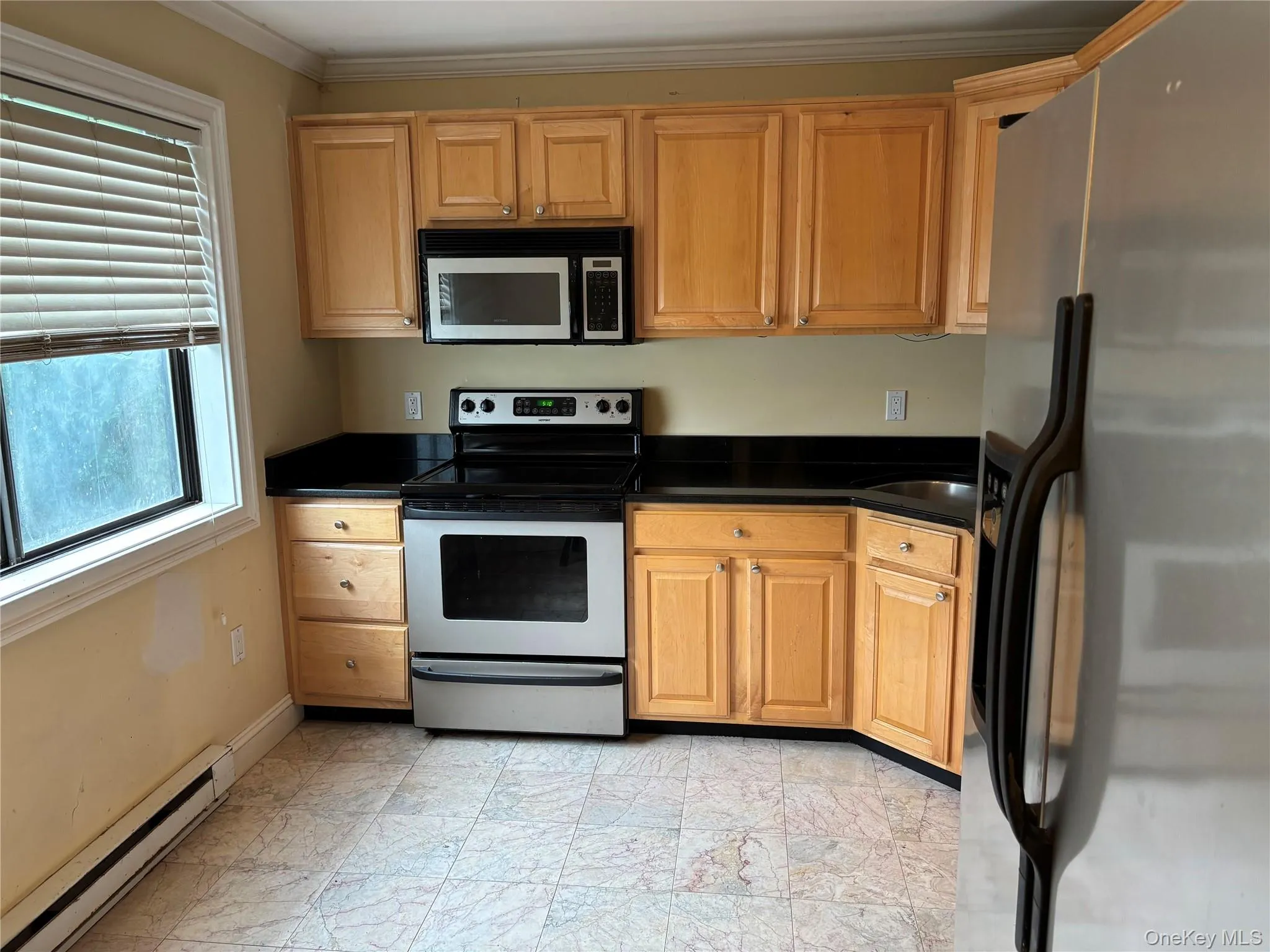Kitchen featuring light wood cabinetry, black countertops, stainless steel appliances, and a window with blinds Kitchen featuring light wood cabinetry, black countertops, stainless steel appliances, and a window with blinds