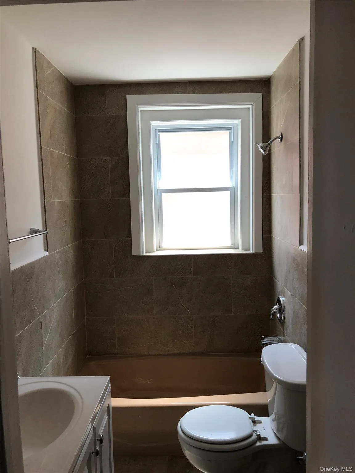 Bathroom featuring a single window above the bathtub, full-height wall tiling, a white vanity with integrated sink, and a white toilet Bathroom featuring a single window above the bathtub, full-height wall tiling, a white vanity with integrated sink, and a white toilet
