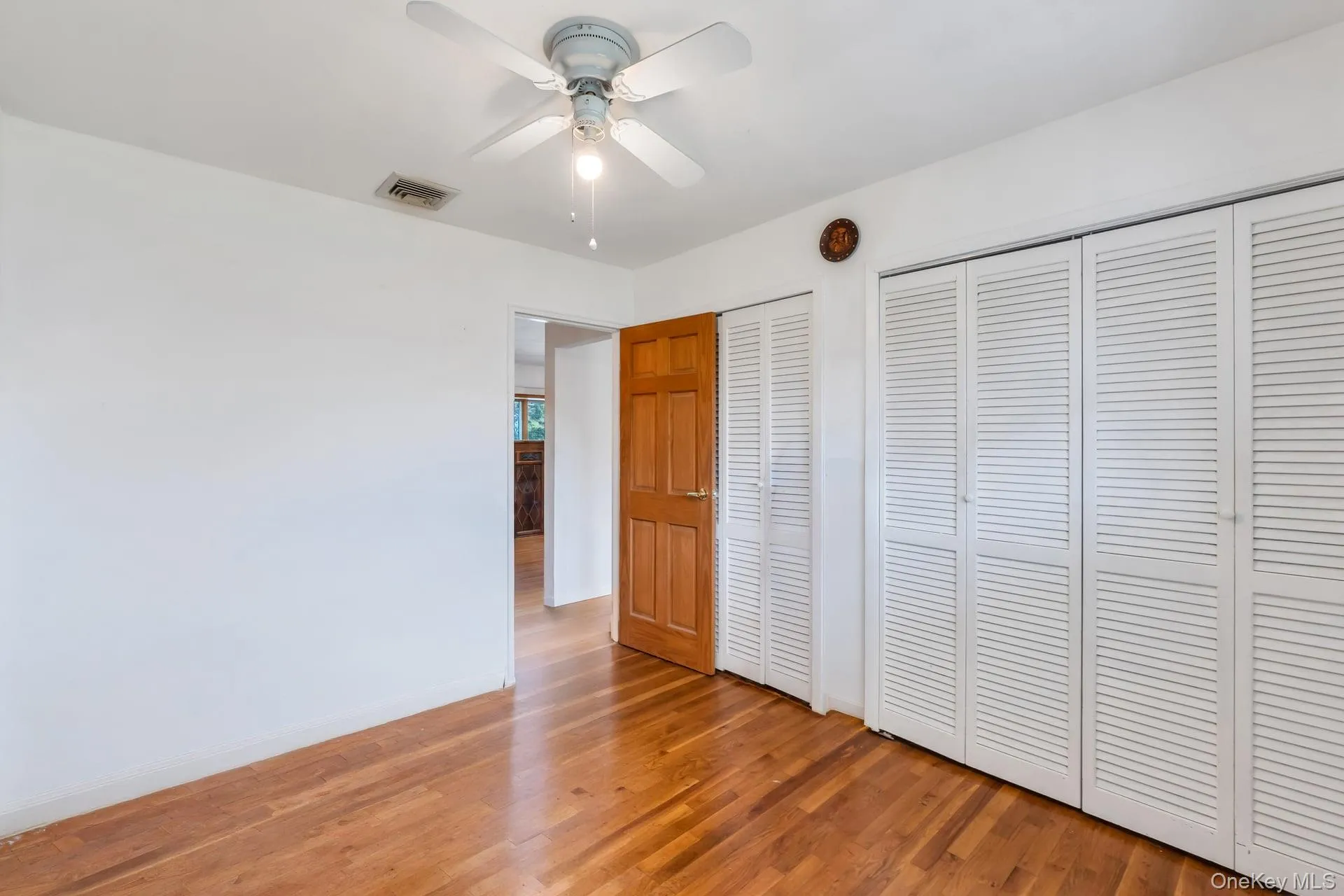 Hardwood flooring throughout, featuring extensive louvered closet doors, a paneled wood door, and a ceiling fan with integrated lighting Hardwood flooring throughout, featuring extensive louvered closet doors, a paneled wood door, and a ceiling fan with integrated lighting
