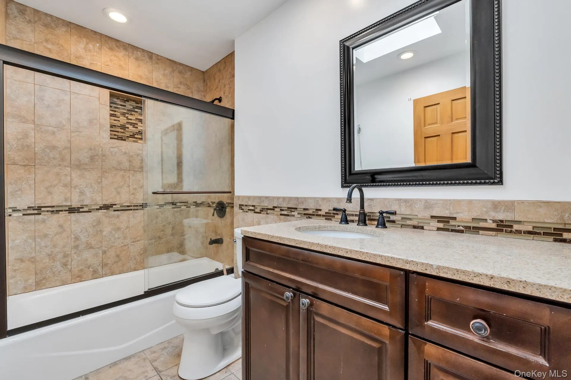 Bathroom featuring a dark wood-finish vanity with a light-toned countertop, a frameless shower with a sliding glass door, mosaic tile accents, and a skylight Bathroom featuring a dark wood-finish vanity with a light-toned countertop, a frameless shower with a sliding glass door, mosaic tile accents, and a skylight