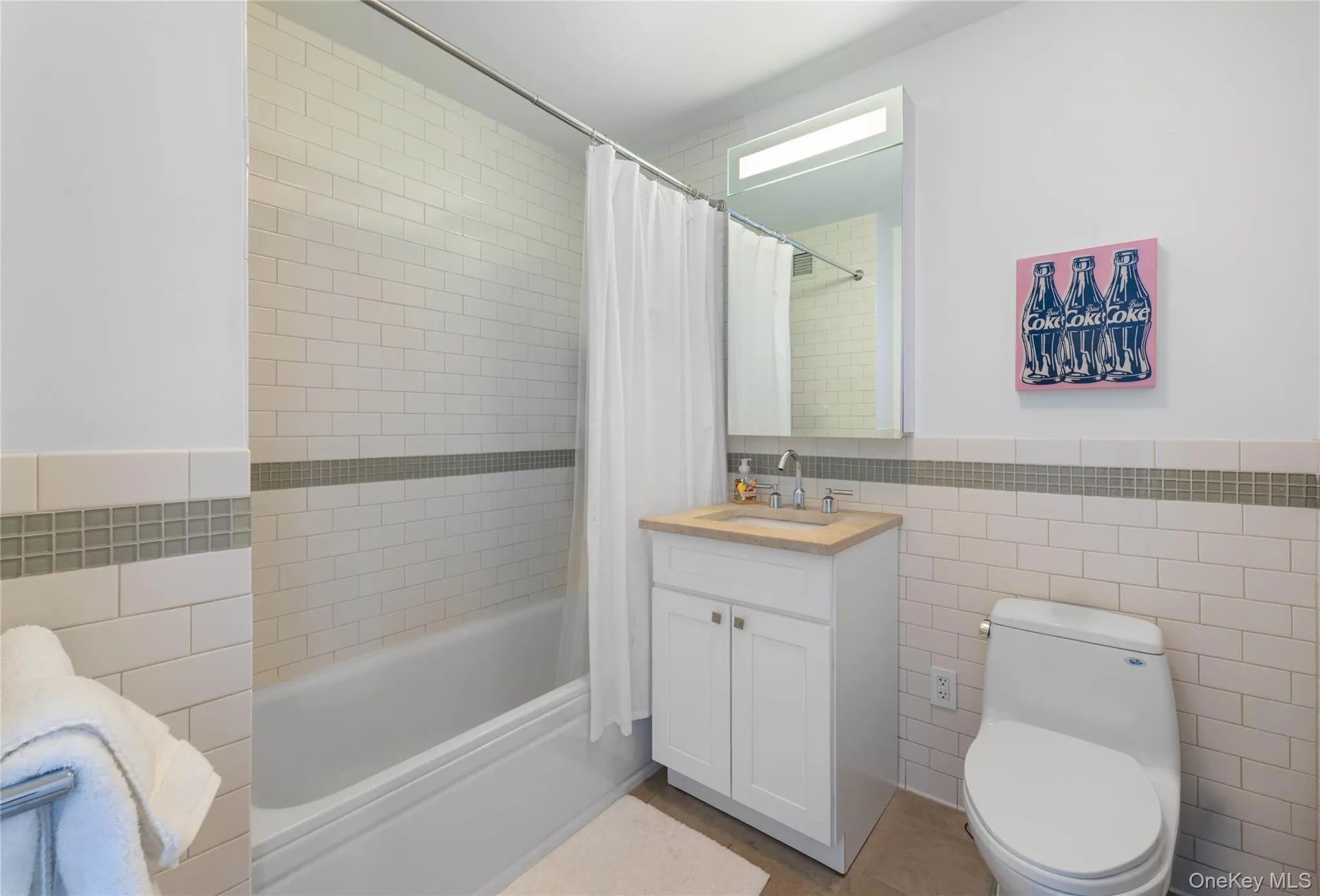 Bathroom featuring a white subway tile surround with a glass mosaic accent band, a white vanity with a light-toned countertop, a rectangular mirror with integrated lighting, and a white toilet Bathroom featuring a white subway tile surround with a glass mosaic accent band, a white vanity with a light-toned countertop, a rectangular mirror with integrated lighting, and a white toilet