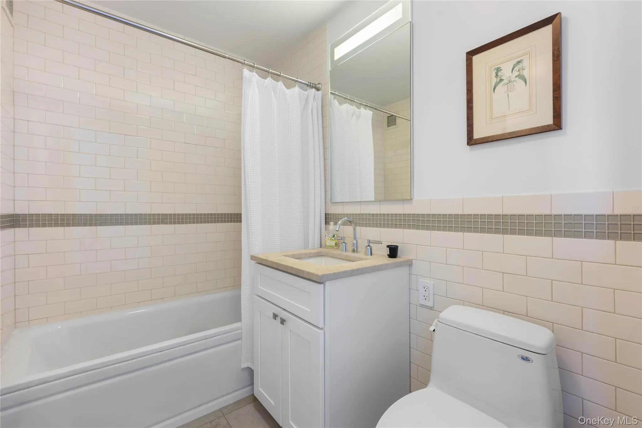 Bathroom featuring a white subway tile surround with a glass mosaic accent band, a white vanity with a light-toned countertop, a rectangular mirror with integrated lighting, and a white bathtub Bathroom featuring a white subway tile surround with a glass mosaic accent band, a white vanity with a light-toned countertop, a rectangular mirror with integrated lighting, and a white bathtub