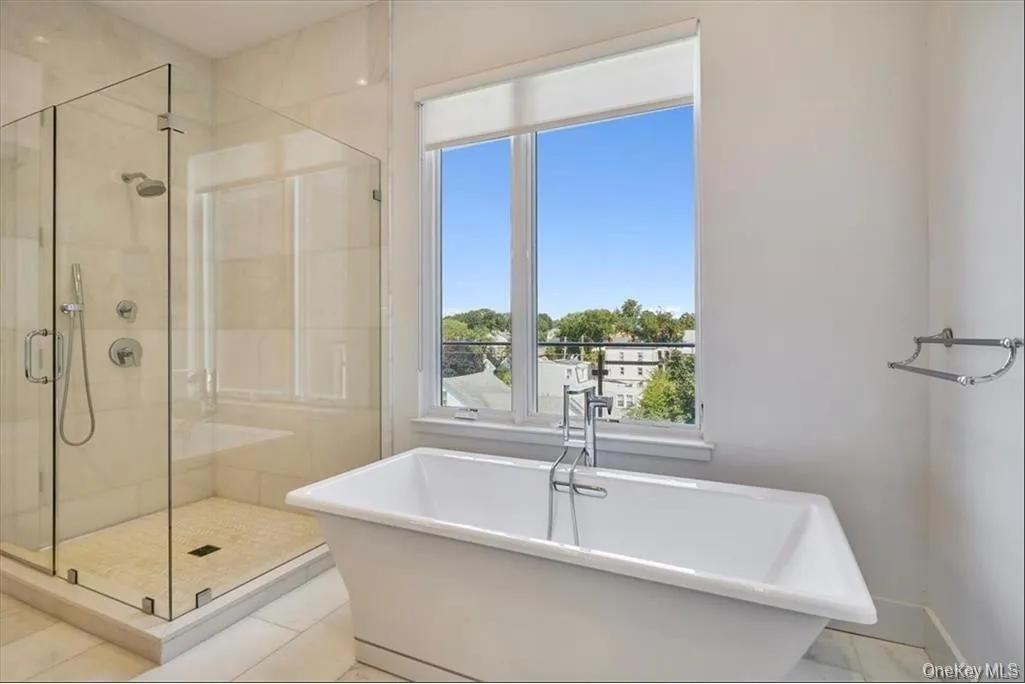 Freestanding soaking tub with chrome fixtures, large window with roller shade, glass-enclosed shower with tiled walls and floor, rain shower head, and hand-held shower sprayer Freestanding soaking tub with chrome fixtures, large window with roller shade, glass-enclosed shower with tiled walls and floor, rain shower head, and hand-held shower sprayer