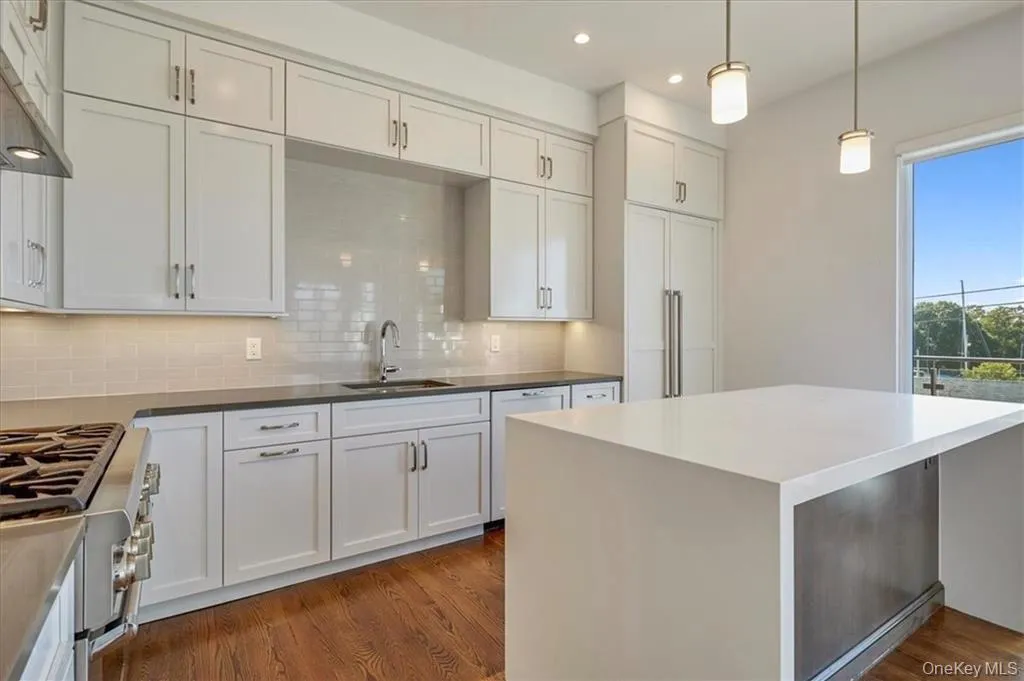 Modern kitchen featuring white shaker cabinetry, stainless steel appliances, a center island with a waterfall countertop, and rich wood-finish flooring Modern kitchen featuring white shaker cabinetry, stainless steel appliances, a center island with a waterfall countertop, and rich wood-finish flooring