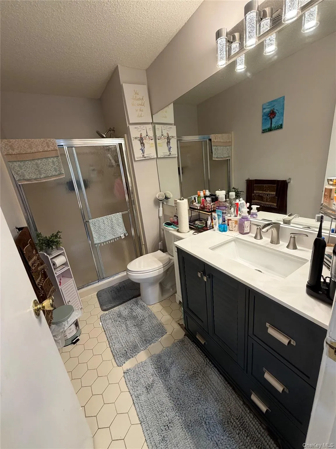 Bathroom featuring a dark vanity with a white countertop and undermount sink, hexagonal floor tiling, a glass-enclosed shower, a wall-mounted light fixture, and a toilet Bathroom featuring a dark vanity with a white countertop and undermount sink, hexagonal floor tiling, a glass-enclosed shower, a wall-mounted light fixture, and a toilet