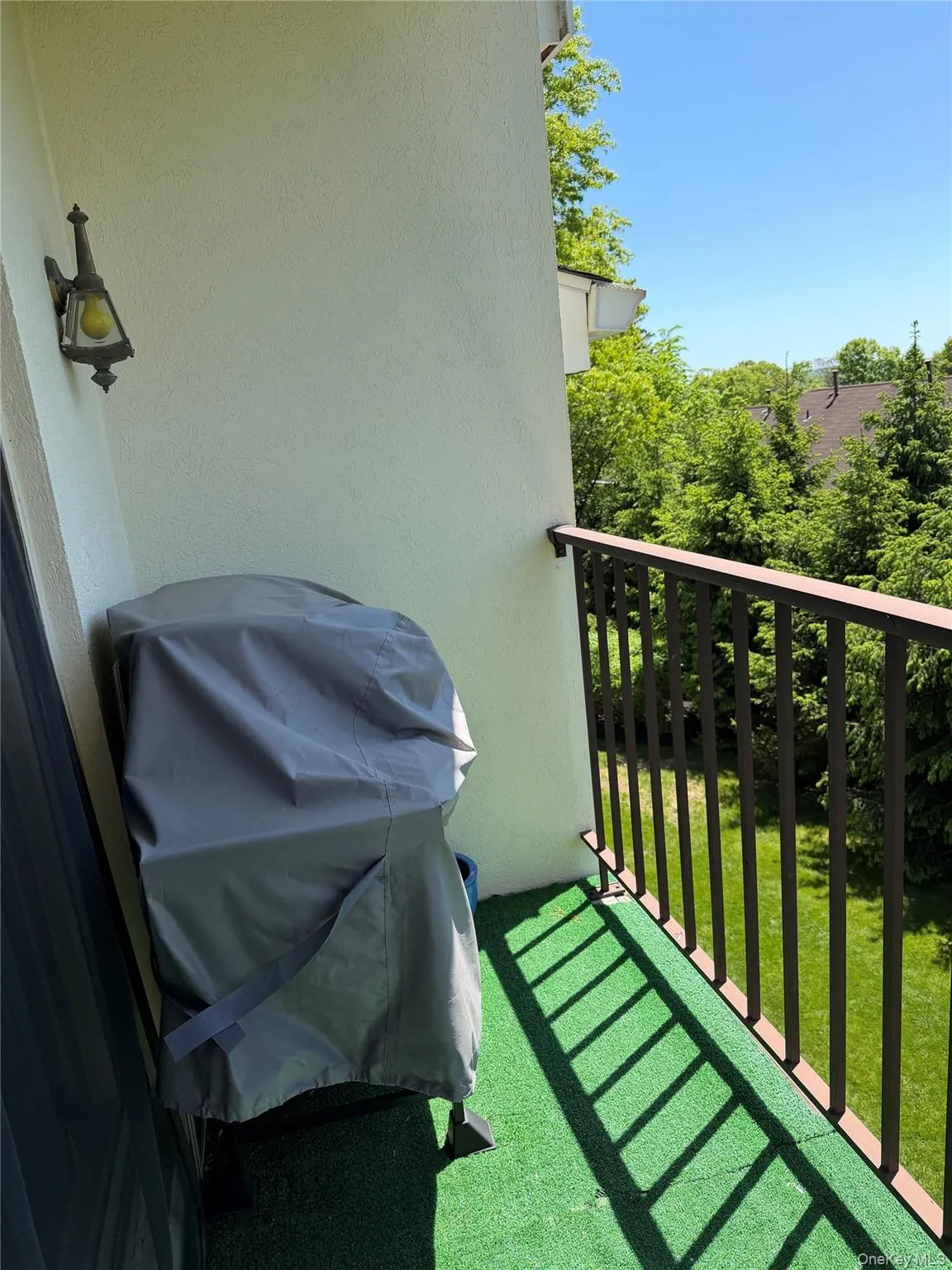 Balcony with a dark brown metal railing, a wall-mounted exterior light fixture, and green turf flooring Balcony with a dark brown metal railing, a wall-mounted exterior light fixture, and green turf flooring