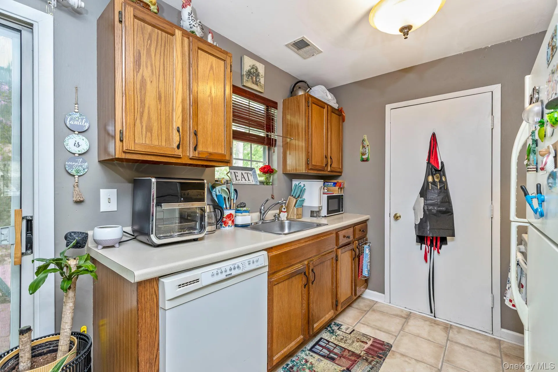 Kitchen featuring wood cabinetry, a light-colored countertop, an undermount sink, a white dishwasher, and tile flooring Kitchen featuring wood cabinetry, a light-colored countertop, an undermount sink, a white dishwasher, and tile flooring