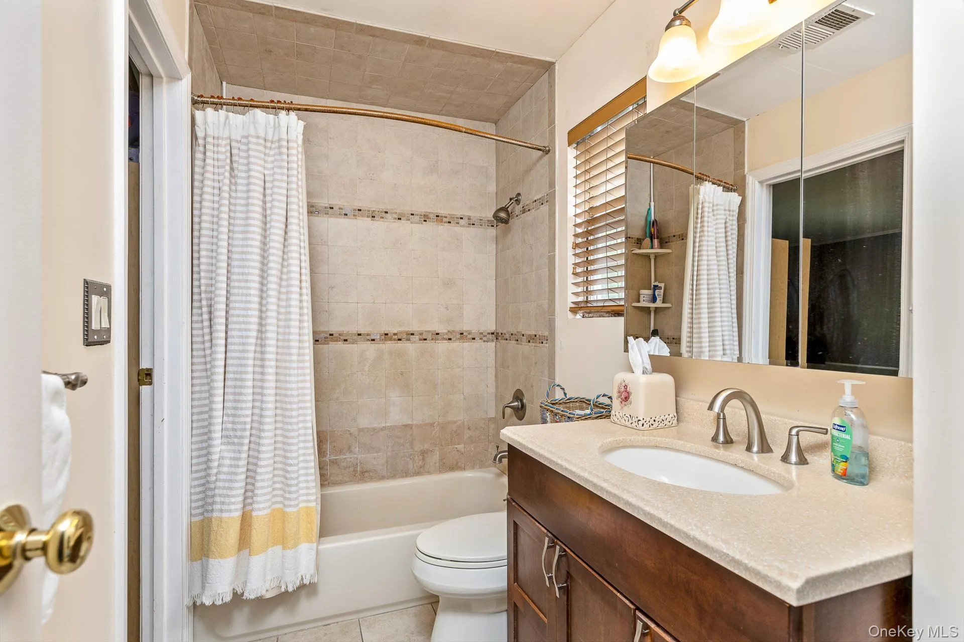 Bathroom featuring a wood-finish vanity with a light-toned countertop, a shower-over-tub combination with tiled surround, and a wall-mounted mirror above the vanity Bathroom featuring a wood-finish vanity with a light-toned countertop, a shower-over-tub combination with tiled surround, and a wall-mounted mirror above the vanity