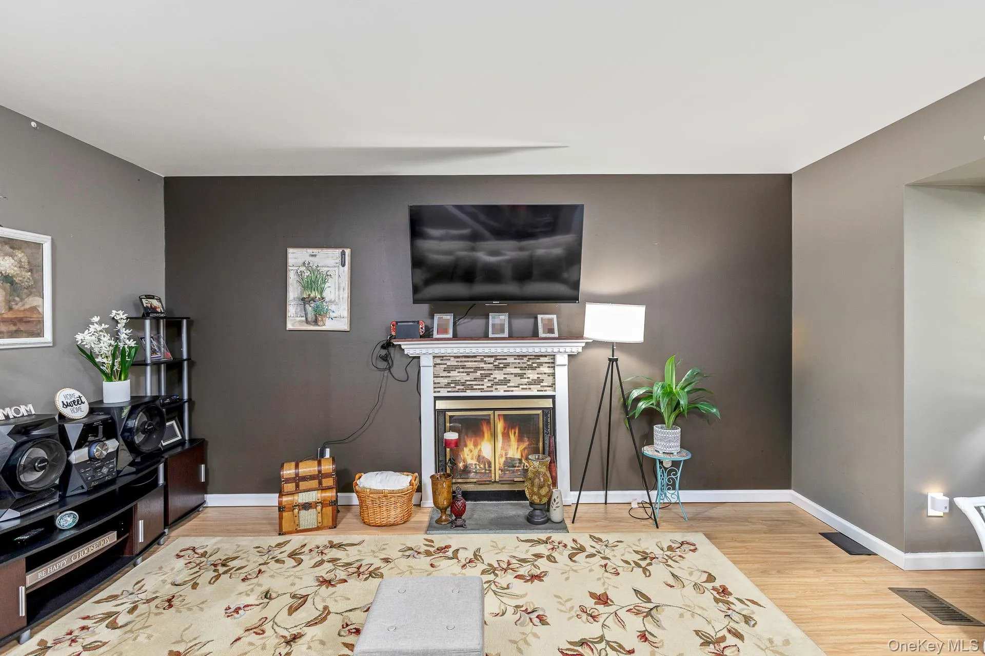 Living area featuring wood-finish flooring, a fireplace with a stacked stone facade and white mantel, and a dark accent wall Living area featuring wood-finish flooring, a fireplace with a stacked stone facade and white mantel, and a dark accent wall