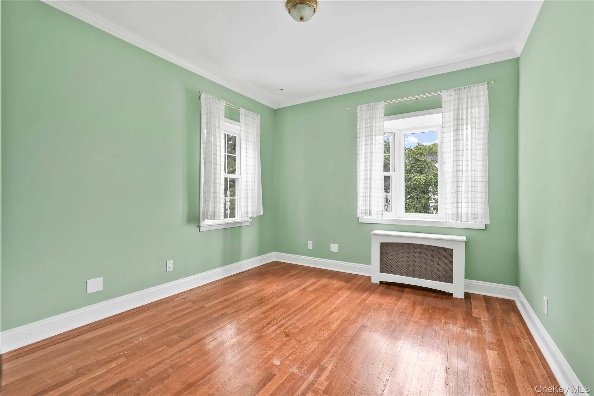 Bedroom 1Room featuring wood flooring, two windows, crown molding, baseboards, and a radiator with cover Bedroom 1Room featuring wood flooring, two windows, crown molding, baseboards, and a radiator with cover