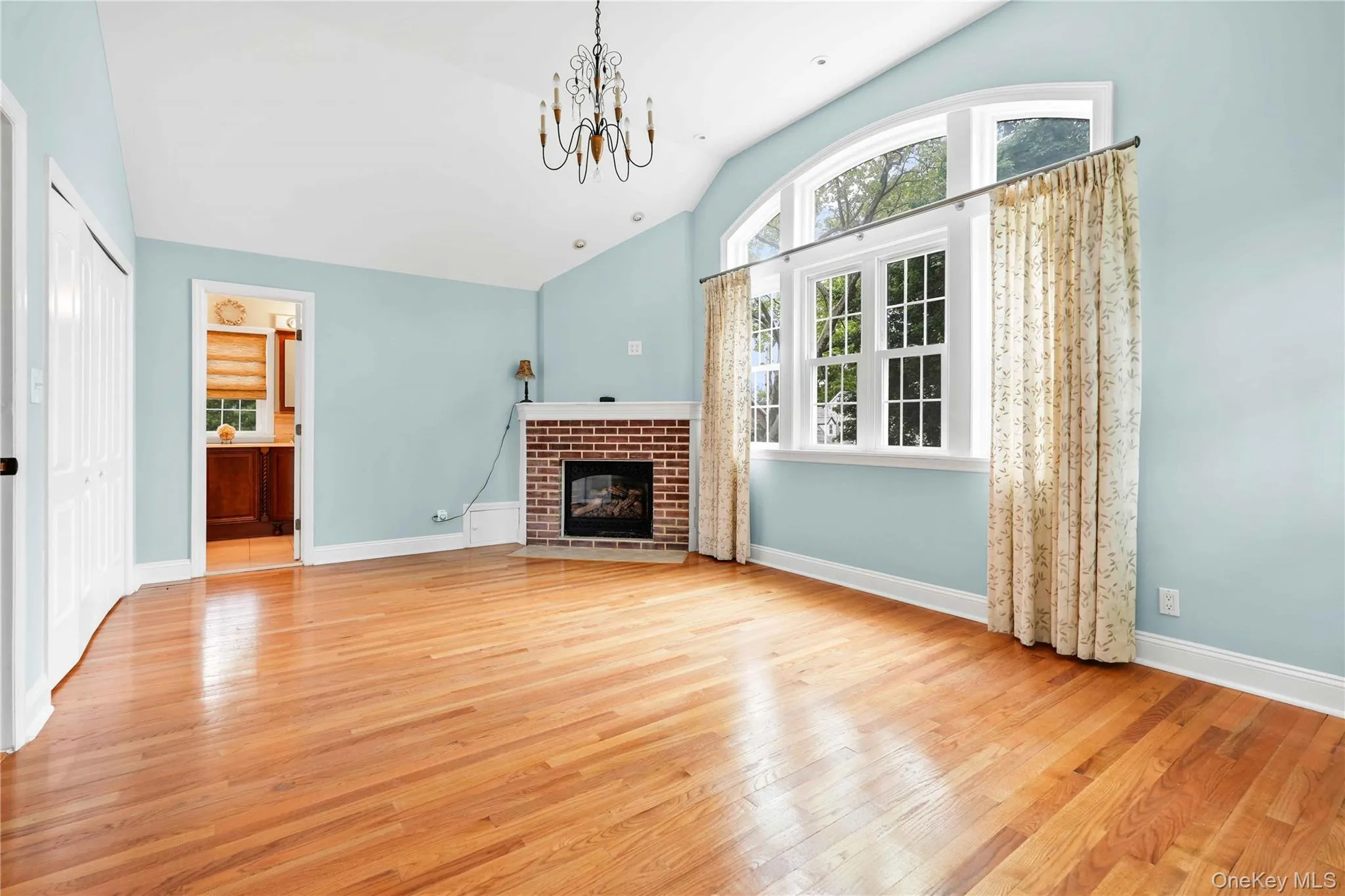 Main Bedroomroom featuring wood-finish flooring, a brick gas fireplace, a large arched window, and a decorative chandelier. Tree views Main Bedroomroom featuring wood-finish flooring, a brick gas fireplace, a large arched window, and a decorative chandelier. Tree views