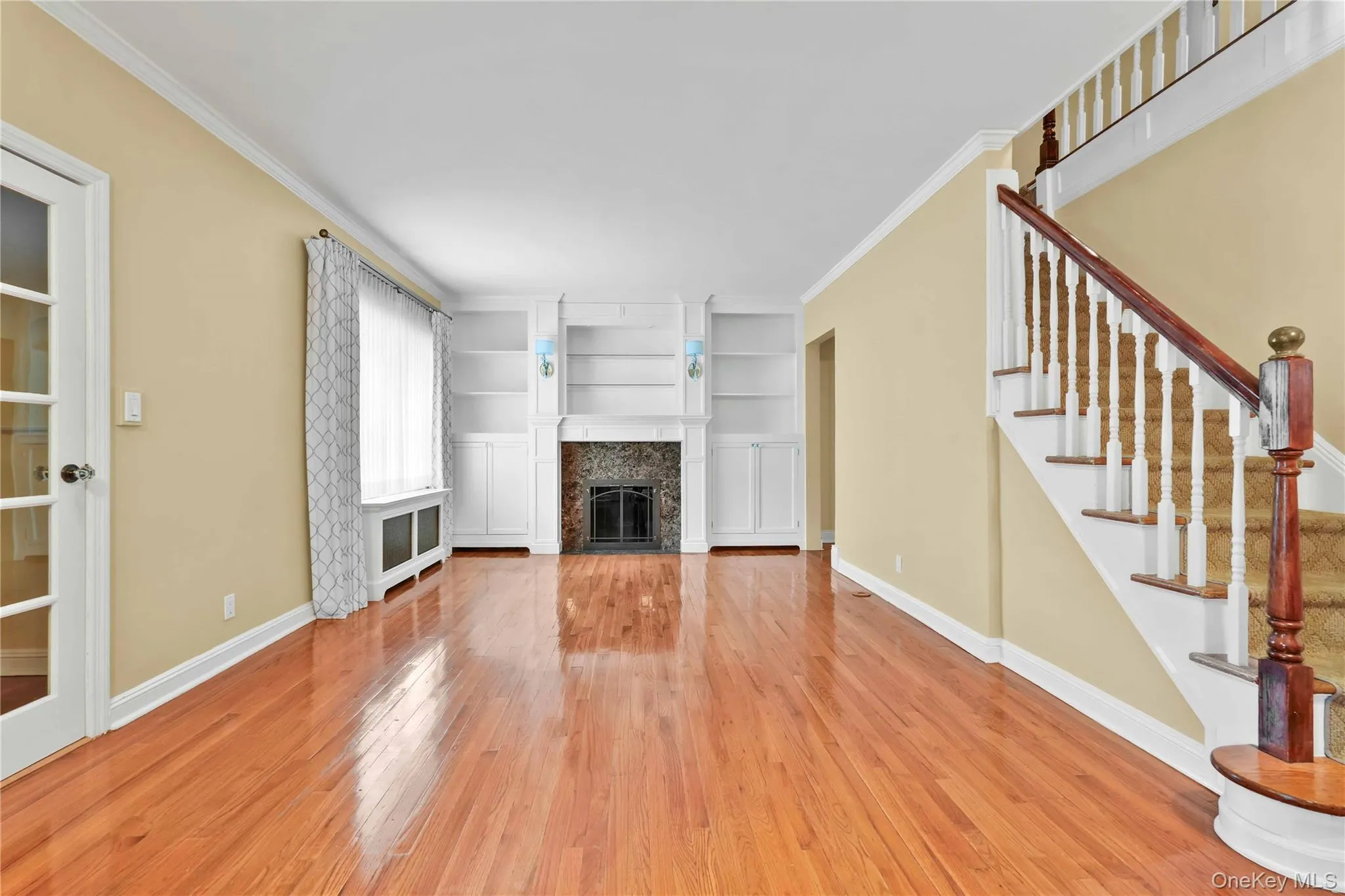Spacious living area featuring polished wood-finish flooring, a central fireplace with an ornate stone surround, and built-in shelving Spacious living area featuring polished wood-finish flooring, a central fireplace with an ornate stone surround, and built-in shelving