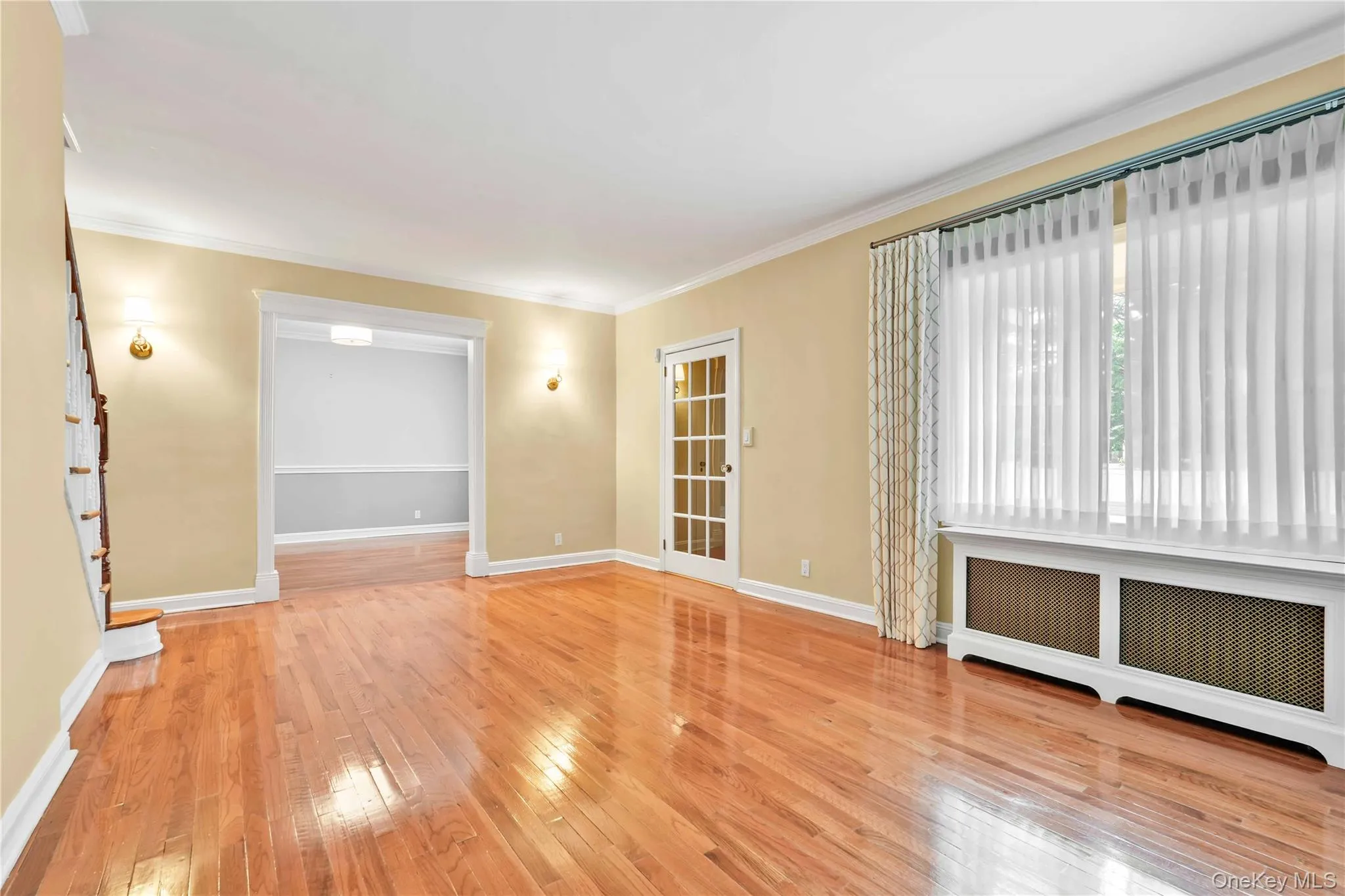 Hardwood flooring throughout, featuring a large picture window with a decorative radiator cover, two wall sconces, and a French door Hardwood flooring throughout, featuring a large picture window with a decorative radiator cover, two wall sconces, and a French door