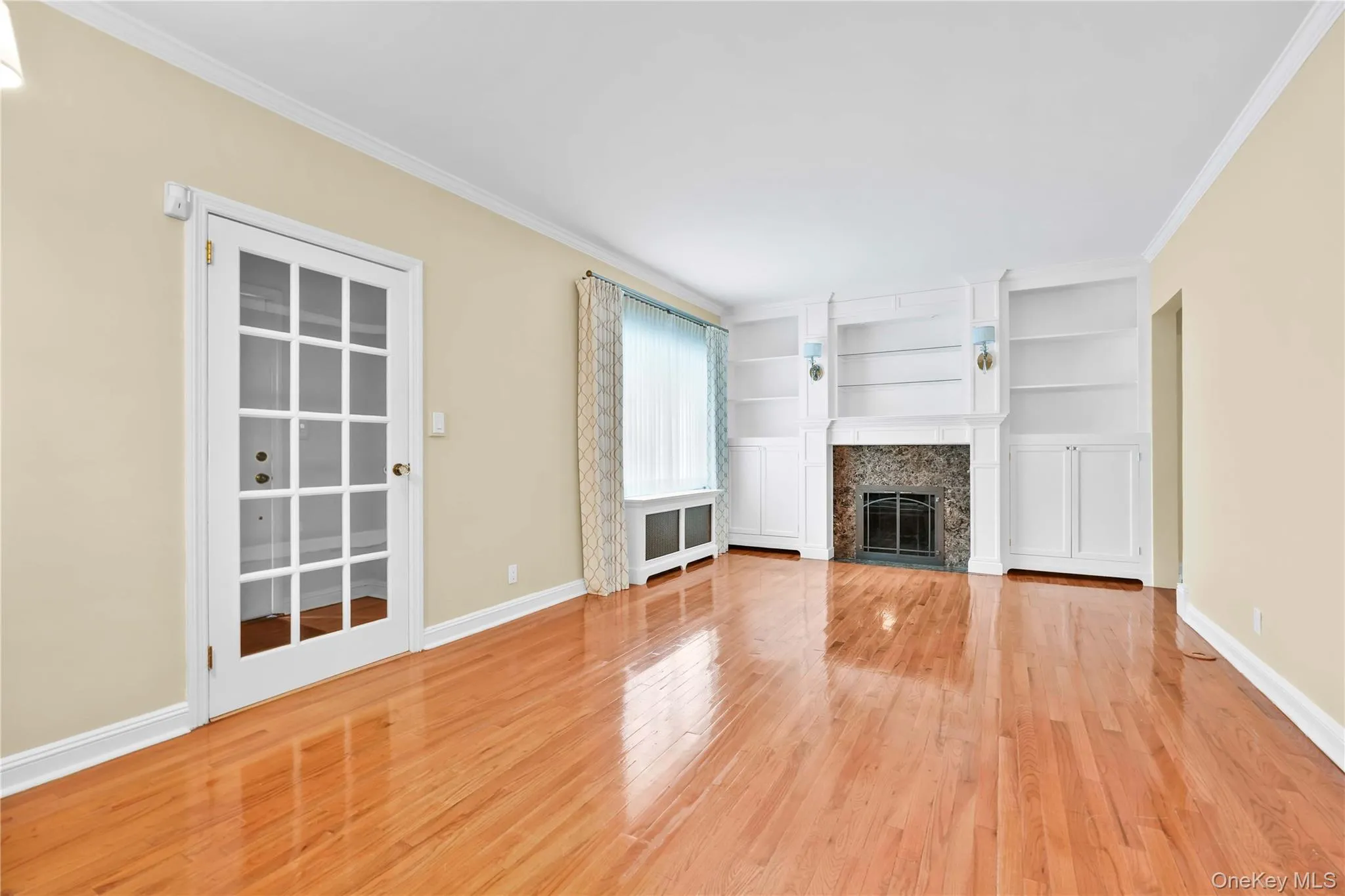 Spacious interior featuring wood-finish flooring, a fireplace with stone surround, custom built-in shelving, a French door, and crown molding Spacious interior featuring wood-finish flooring, a fireplace with stone surround, custom built-in shelving, a French door, and crown molding
