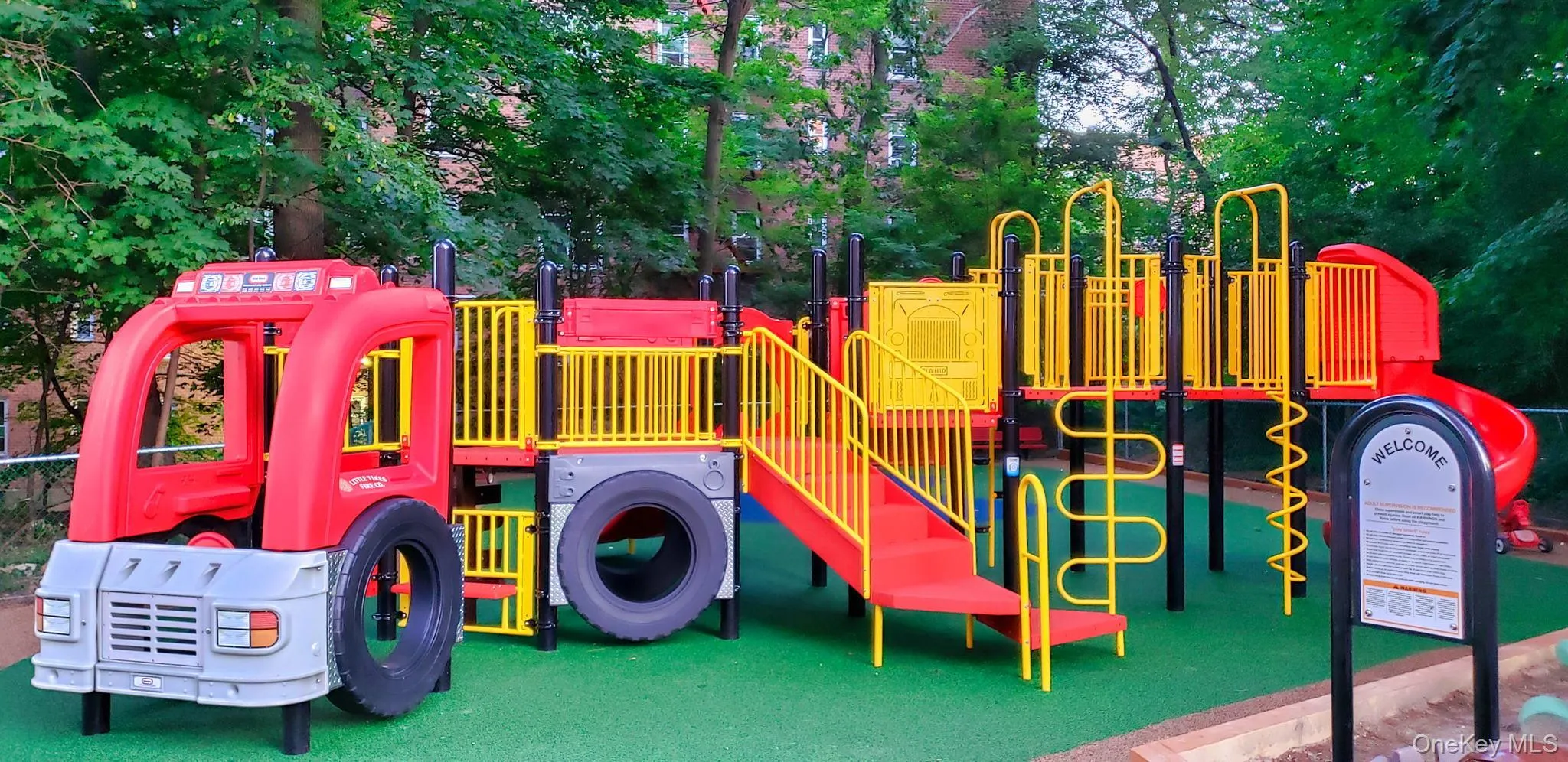 Outdoor play structure featuring a fire truck facade, multiple slides, climbing elements, and a soft-surface green ground Outdoor play structure featuring a fire truck facade, multiple slides, climbing elements, and a soft-surface green ground