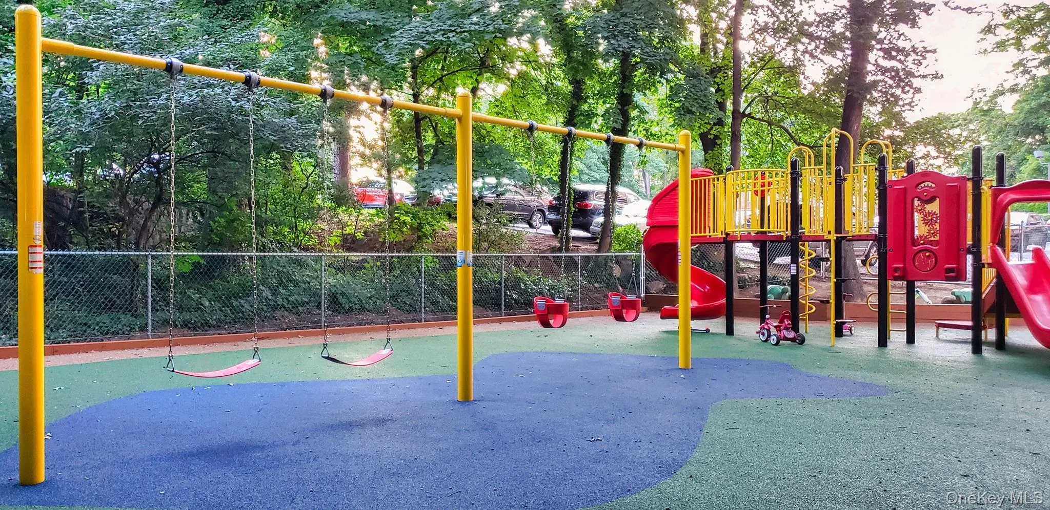 Outdoor recreation area featuring a swing set with two swings, a multi-level play structure with red slides, and a soft-surface ground cover in blue and green Outdoor recreation area featuring a swing set with two swings, a multi-level play structure with red slides, and a soft-surface ground cover in blue and green