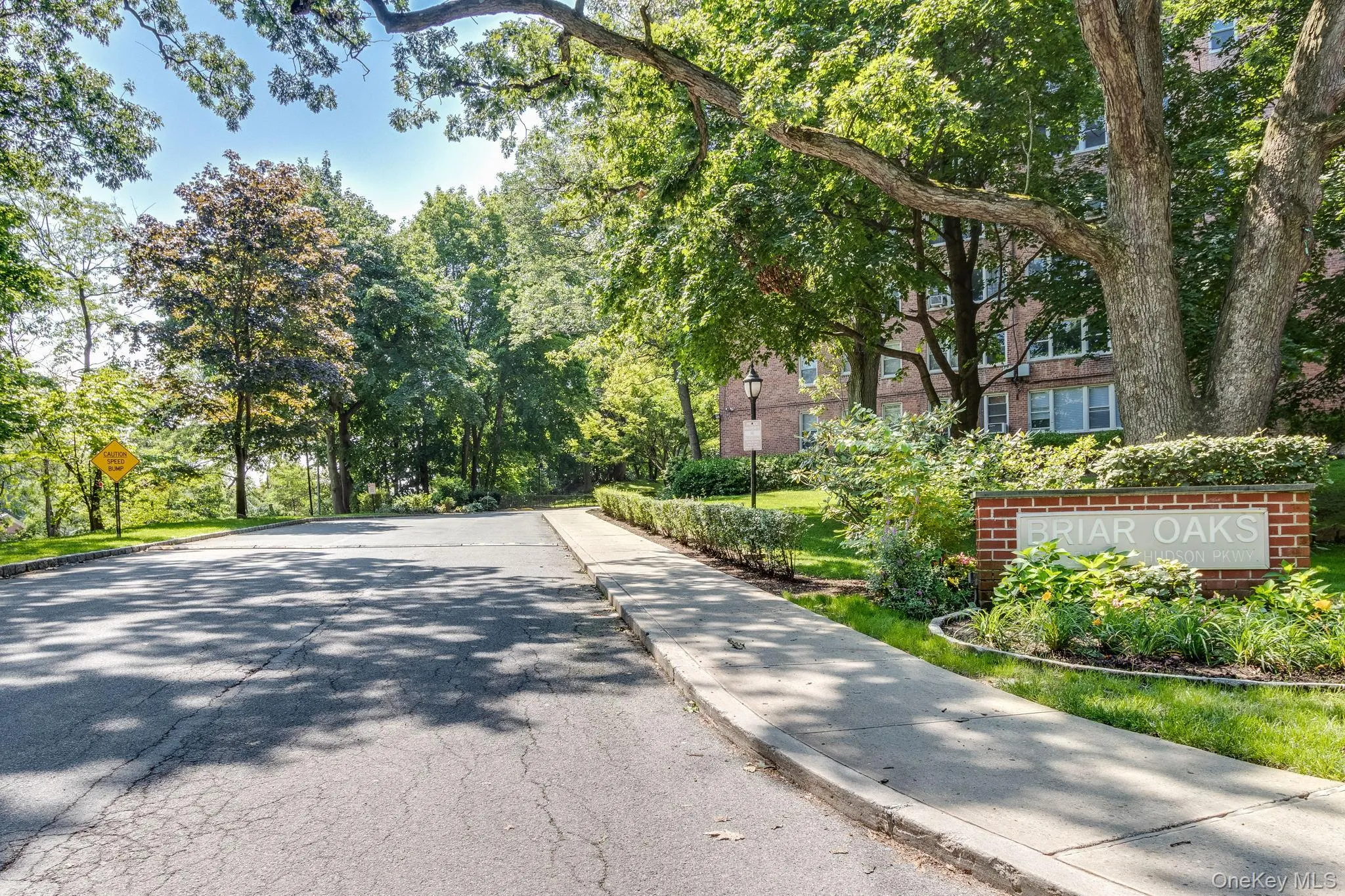 Paved residential roadway with a concrete sidewalk and brick entrance sign for "BRIAR OAKS." Mature trees provide natural canopy, complemented by manicured shrubbery and green lawns Paved residential roadway with a concrete sidewalk and brick entrance sign for "BRIAR OAKS." Mature trees provide natural canopy, complemented by manicured shrubbery and green lawns
