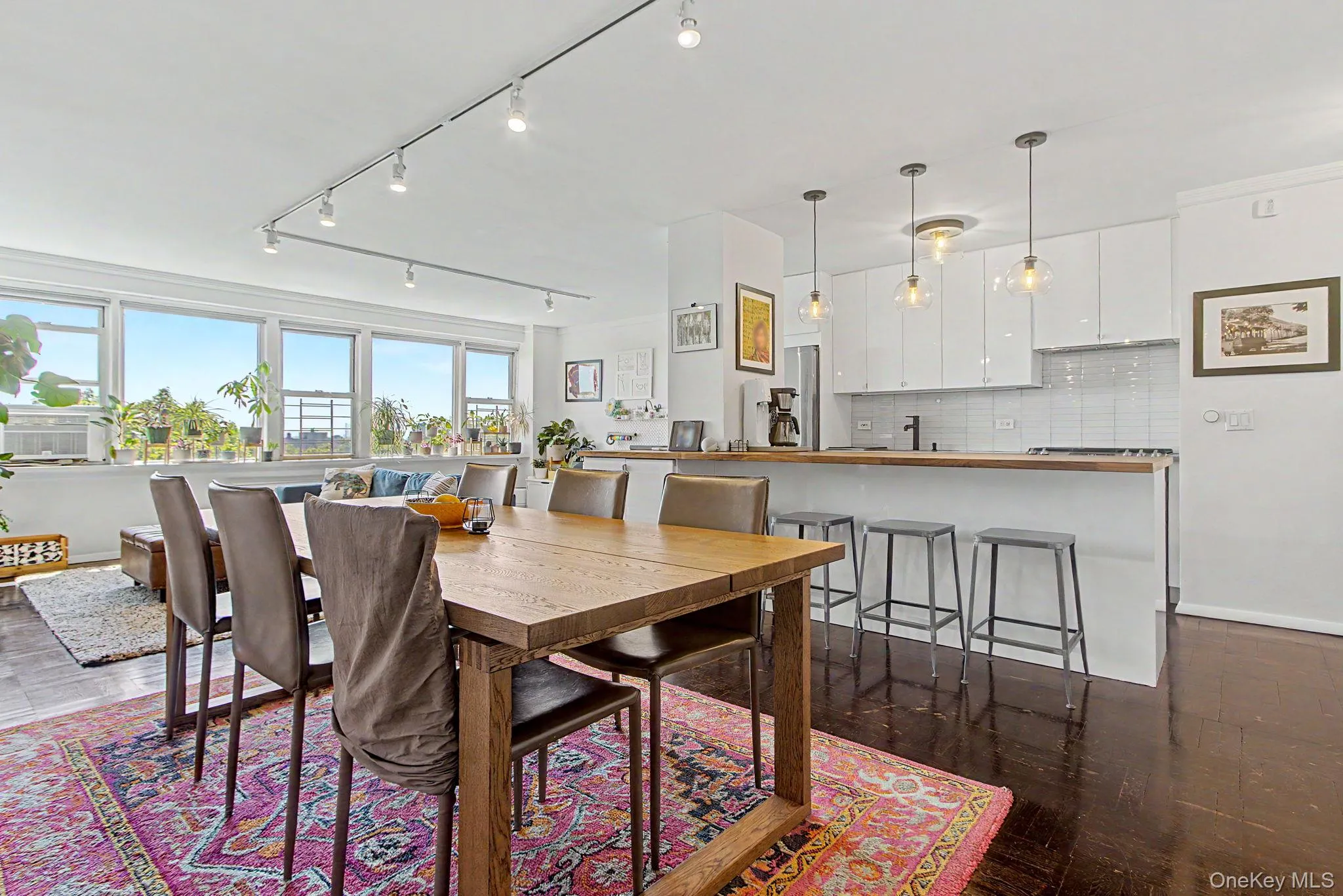 Open-concept living space featuring expansive windows, dark wood-finish flooring, a contemporary kitchen with white cabinetry and a breakfast bar, overhead track lighting, and pendant lighting fixtures Open-concept living space featuring expansive windows, dark wood-finish flooring, a contemporary kitchen with white cabinetry and a breakfast bar, overhead track lighting, and pendant lighting fixtures