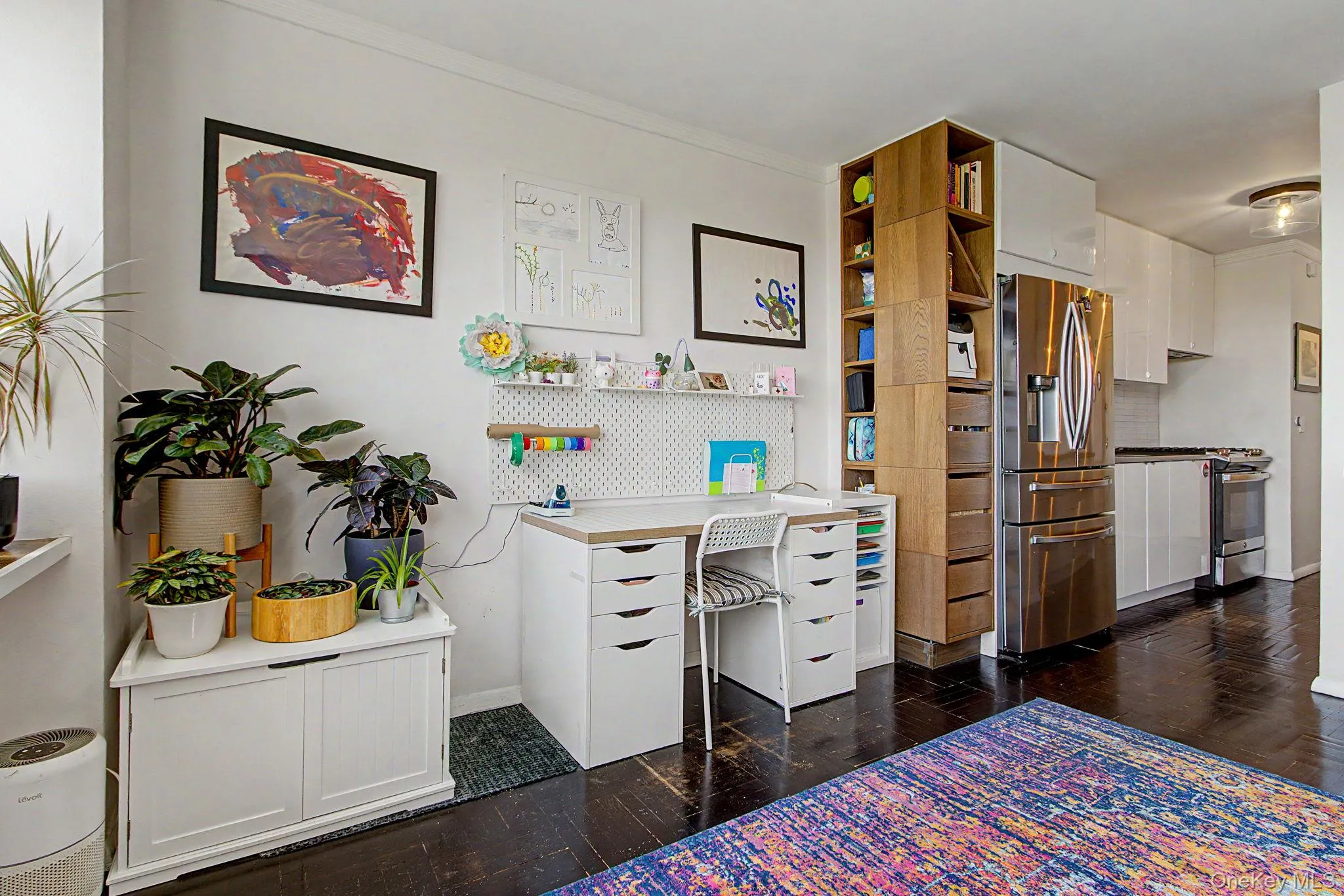Integrated desk area with a pegboard organizer, dark wood-finish flooring, white kitchen cabinetry, stainless steel refrigerator, and built-in wood-finish shelving Integrated desk area with a pegboard organizer, dark wood-finish flooring, white kitchen cabinetry, stainless steel refrigerator, and built-in wood-finish shelving